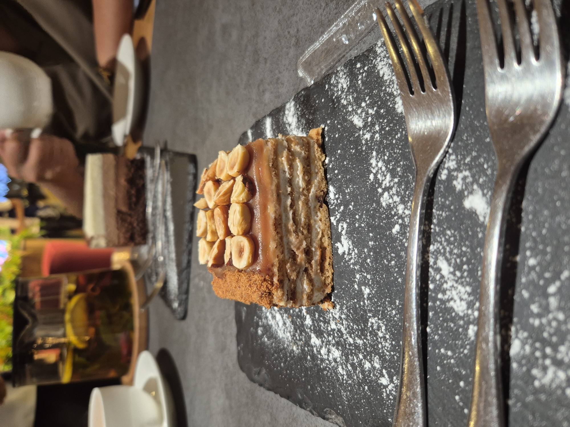 The image depicts a slice of nut-topped cake on a black slate plate dusted with powdered sugar, along with two forks. Another dessert and a teapot with lemon are visible in the background. The scene appears to be in a restaurant or cafe in Kokshetau, Kazakhstan, based on contextual awareness. A person's hand is visible in the background near another dessert plate.