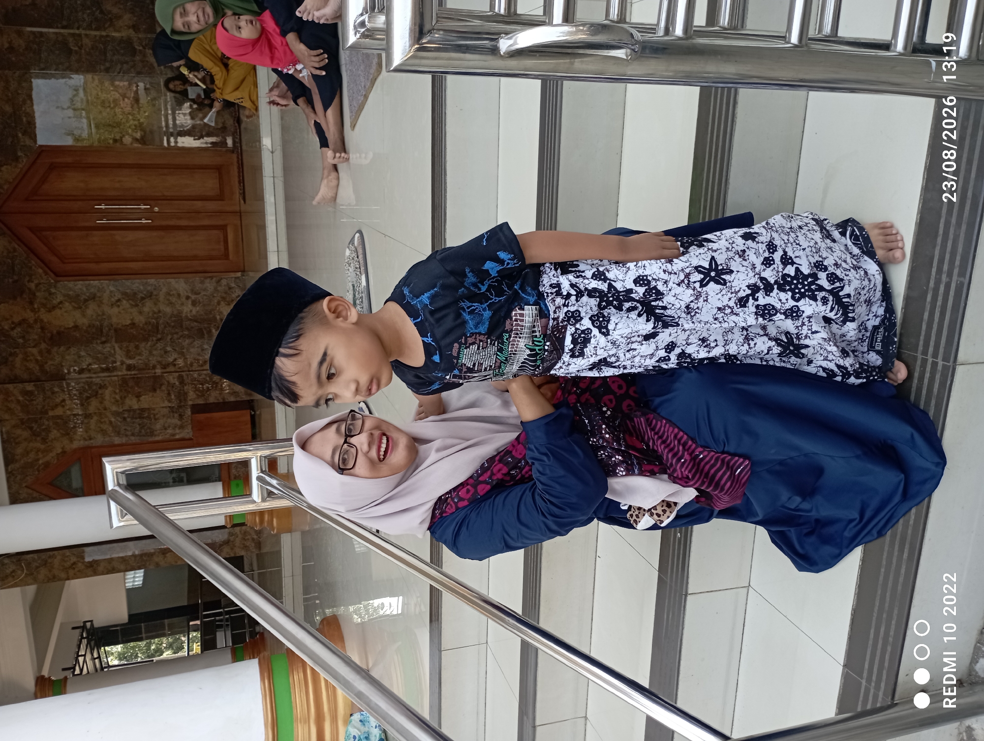 In Dukuhseti, Indonesia, a woman wearing a navy blue hijab and dress, with glasses and a patterned scarf, kneels on a tiled staircase. Beside her, a young boy in a black cap, black t-shirt with a blue design, and a white sarong with black patterns, stands facing slightly away from the camera. They are on a staircase with stainless steel railings, with a large wooden door and tiled walls in the background. Other people are visible in the background, some seated on the floor. The timestamp "23/08/2026 13:19" is visible in the lower right corner, along with "REDMI 10 2022" in the lower left.