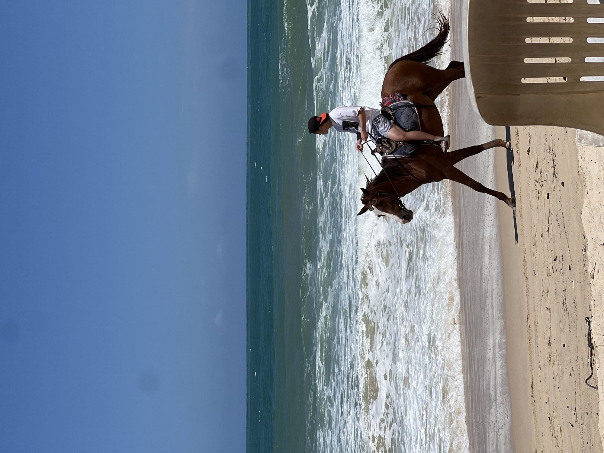 A person rides a brown horse along the sandy shore of a beach, with the turquoise ocean and white waves in the background under a clear blue sky. The rider, wearing a white t-shirt, striped shorts, and a baseball cap, is positioned on the horse's back, holding the reins. The horse is walking with its front left leg extended forward. The beach sand is light brown and appears wet near the water's edge. In the foreground, a light-colored plastic chair is partially visible, suggesting a recreational setting. The scene depicts a serene moment of a person enjoying a horseback ride by the sea. This location is within the city of Choeng Thale, Thailand.