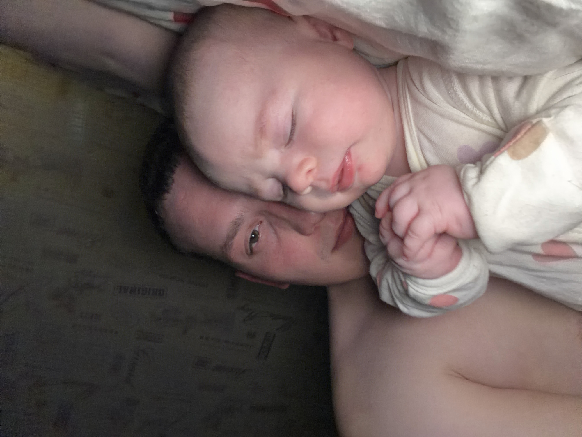 A close-up, angled, indoor photograph shows a baby sleeping on an adult. The baby has a pale complexion and is wearing a cream-colored shirt with pink polka dots. The adult is laying on their back with the baby resting on their chest. The adult has dark hair, fair skin, and visible stubble. The location is identifiable as Chaikovsky, Russia. A corner of the photograph shows a dark surface with what appears to be faded text; "ORIGINAL" is discernable.