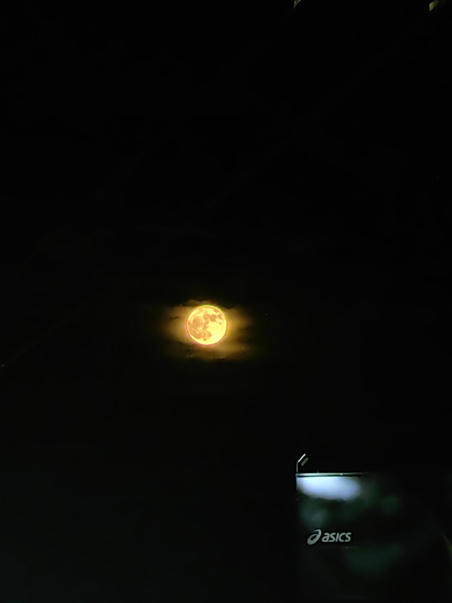 The image shows a bright, orange moon set against a dark, night sky. In the lower right corner, the "Asics" logo is visible, suggesting the scene is located near or associated with Asics branding. The location is unspecified but implied to be an urban or built environment due to the presence of the logo.