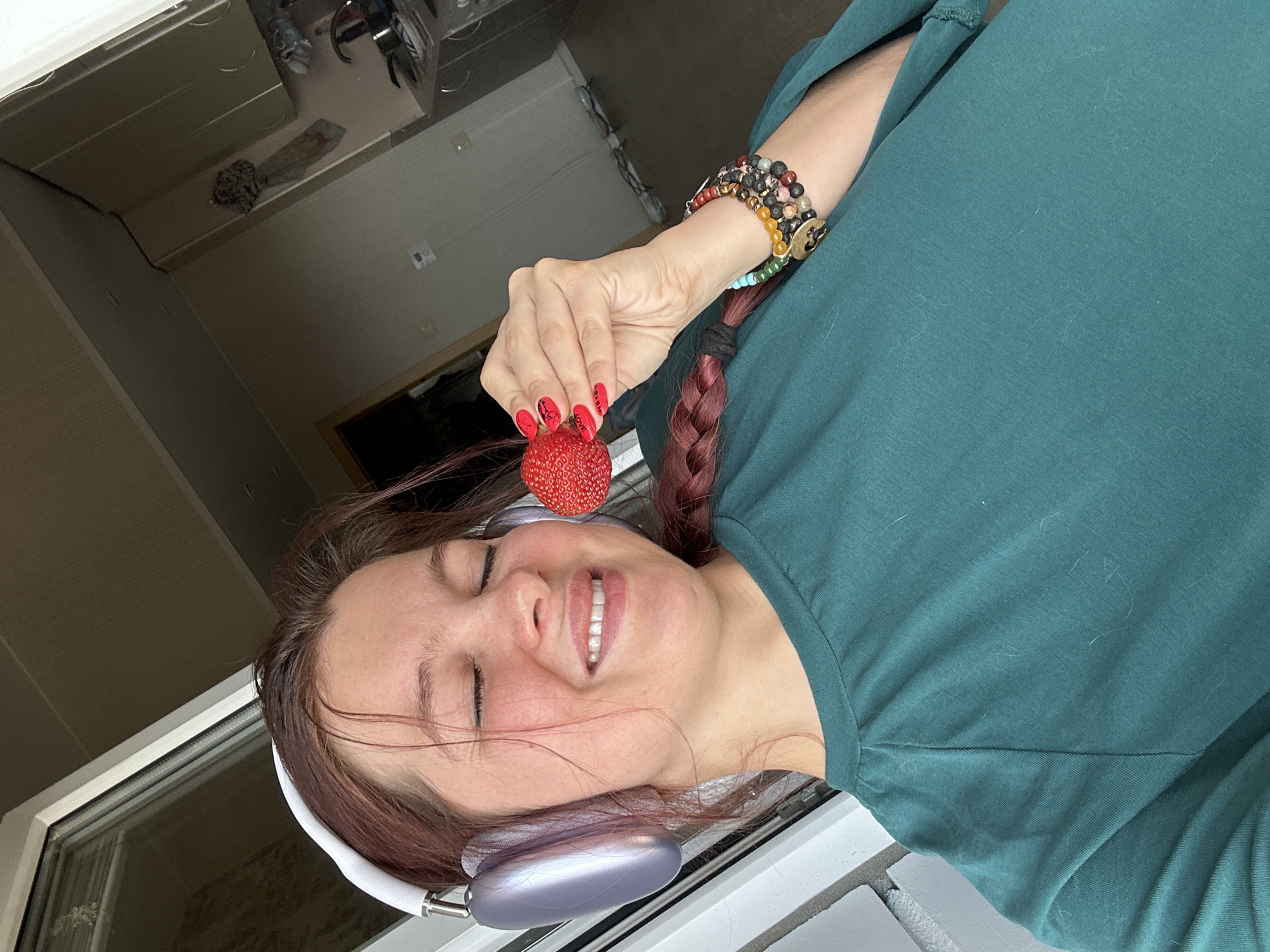 A young woman with auburn hair, wearing headphones and a green t-shirt, is holding a strawberry up to her face. Her eyes are closed and she is smiling. She is wearing bracelets on her left wrist. The background appears to be a domestic interior, possibly a kitchen, with cabinets and appliances visible. The lighting suggests it is daytime. No specific location in Vladimir, Russia can be determined from the image.