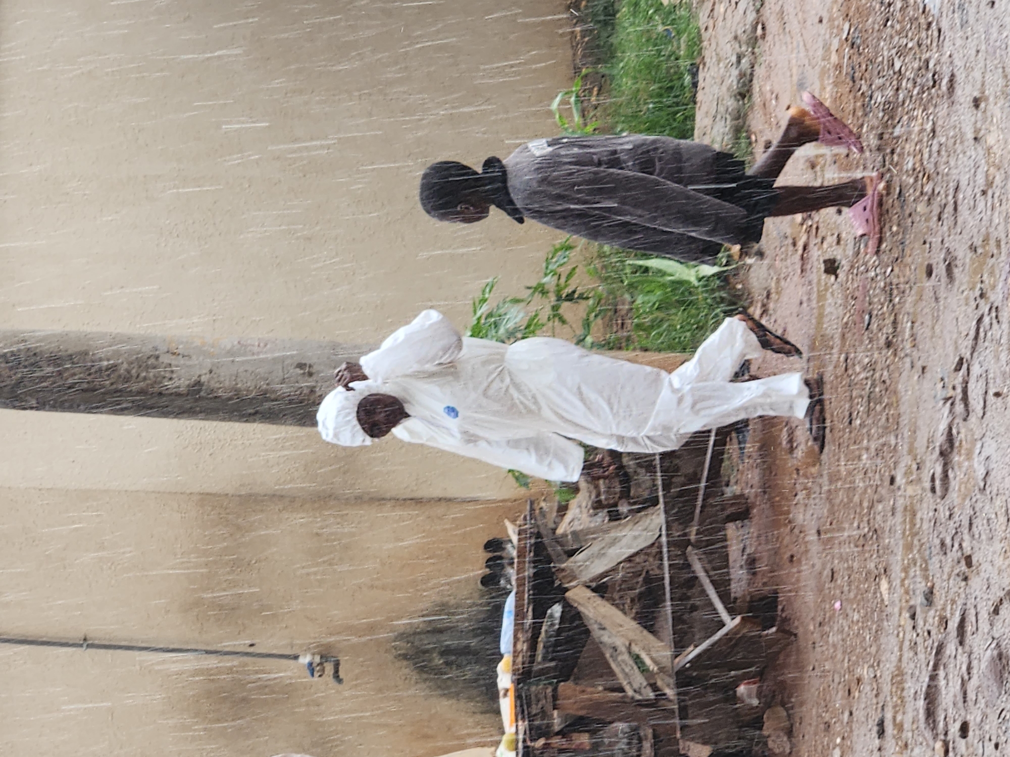 In the Sabon Tasha area of Nigeria, during a heavy downpour, two individuals are captured walking outdoors. On the left, a person is dressed in a white full-body protective suit, possibly a hazmat suit, with a hood pulled over their head. They are walking towards the right of the frame. To their right and slightly ahead, a child is walking in the opposite direction, towards the left of the frame. The child is wearing a dark-colored jacket and shorts, with pink sandals on their feet. The ground is wet and muddy, with visible puddles and scattered debris. Behind the protective-suited individual, a wooden structure, possibly a makeshift stall or table, is partially visible, covered with various items, including what appear to be containers and other objects. A utility pipe with a faucet is mounted on a textured wall in the background, which is also streaked with rain. A thick, rough-textured pole stands between the two individuals, further back in the scene. The overall environment suggests an outdoor, possibly urban or semi-urban setting experiencing inclement weather.