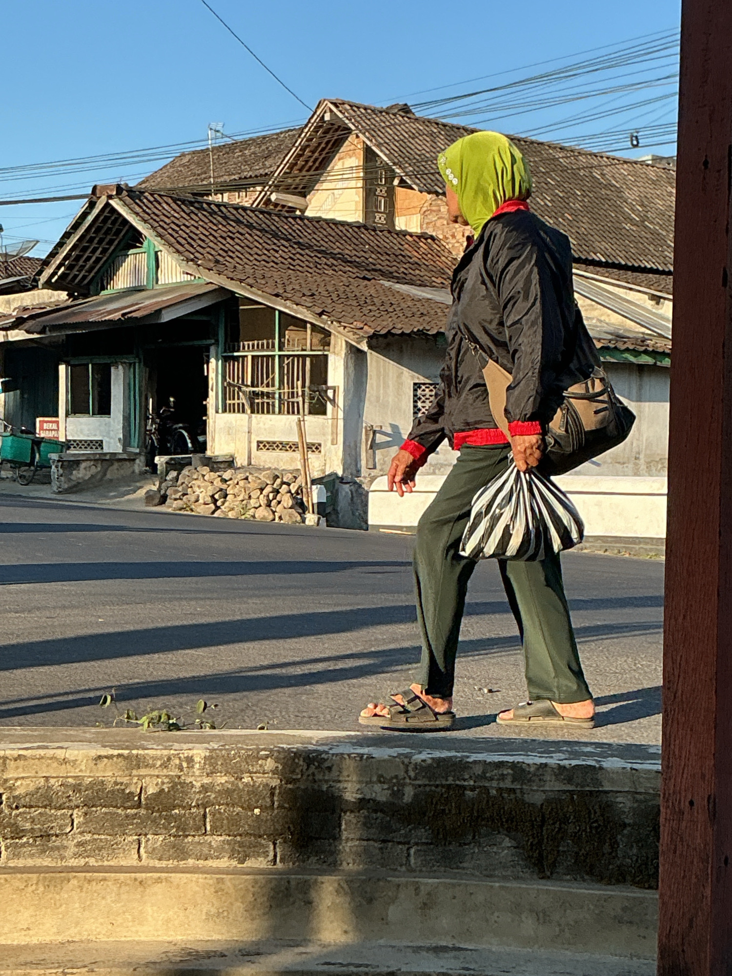 A person wearing a bright green hijab, a black jacket with red cuffs and collar, and dark green pants walks on a paved path next to a street in Kebonrejo, Indonesia. They are carrying a striped bag in their right hand and a messenger bag over their left shoulder. The person is walking away from the camera, towards the right of the frame. Behind them, traditional Indonesian houses with tiled roofs are visible, with some electrical wires running overhead. The ground is a mix of pavement and some overgrown plants. The lighting suggests it is late afternoon or early morning, with long shadows cast on the street.