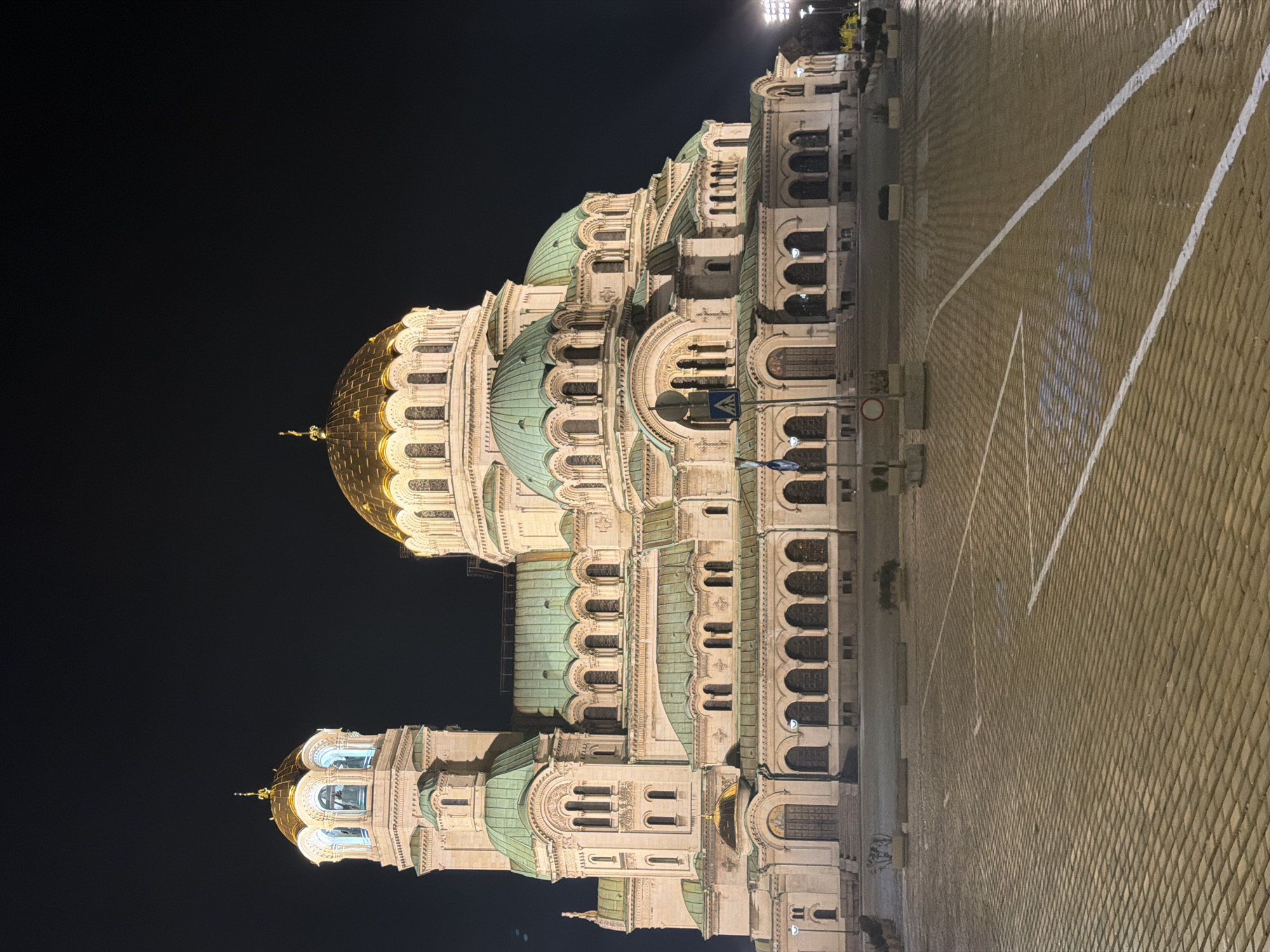 The image shows the Alexander Nevsky Cathedral in Sofia, Bulgaria at night. The cathedral, a large building with multiple domes, is lit up, contrasting with the dark sky. The domes are a mix of gold and green. The foreground is a paved area with white lines marking a court, possibly for a game. No people are visible in the scene. The environment is urban, evidenced by the street lights and the architecture of the cathedral.