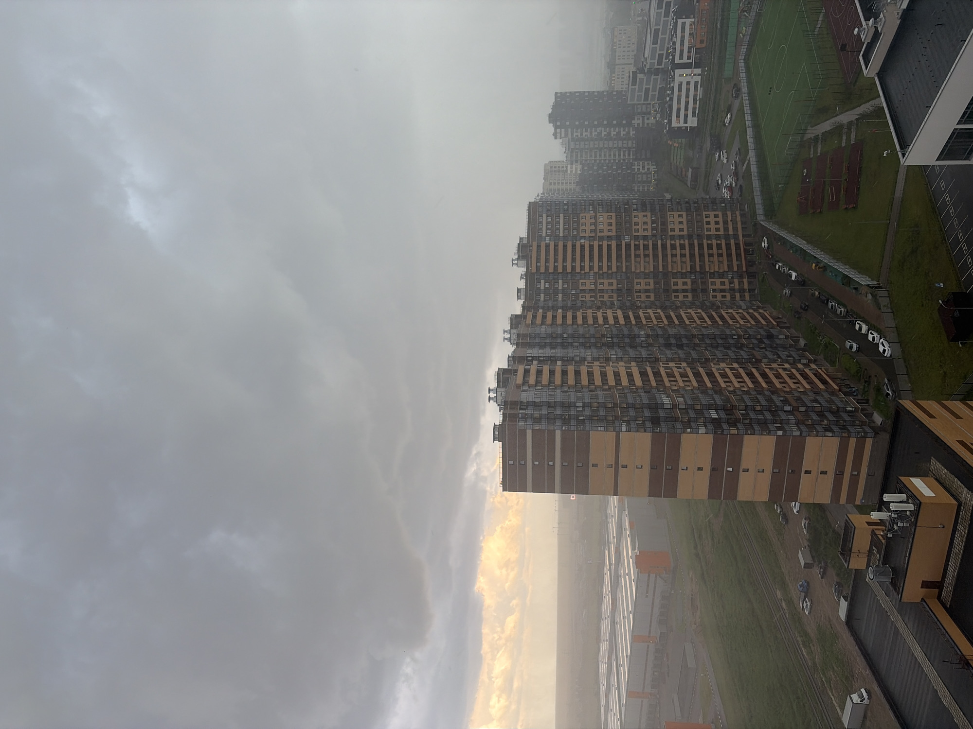 This image captures an aerial view of a residential and industrial area in Saint Petersburg, Russia, likely during an overcast or rainy day. The sky is dominated by heavy, gray clouds with a patch of brighter, yellowish light breaking through on the left, suggesting a sunrise or sunset behind the clouds.
In the foreground and mid-ground, several tall, modern apartment buildings are visible. One prominent building is a multi-story structure with a distinctive facade of horizontal bands of brown and beige. Other apartment buildings are also present, some appearing darker and more distant, blending into the misty atmosphere.
To the left of the apartment buildings, a large industrial complex with multiple long, low-rise buildings, likely warehouses or factories, stretches across the landscape. These buildings have orange and gray accents. A green field, possibly a sports field or park, is situated between some of the residential buildings.
The ground level reveals a network of roads and parking areas with several cars parked. Some vehicles, including vans and cars, are also seen on the roads. The ground appears damp, indicating recent or ongoing precipitation. There are no discernible people in the image. The overall atmosphere is somewhat muted and diffused due to the weather conditions.