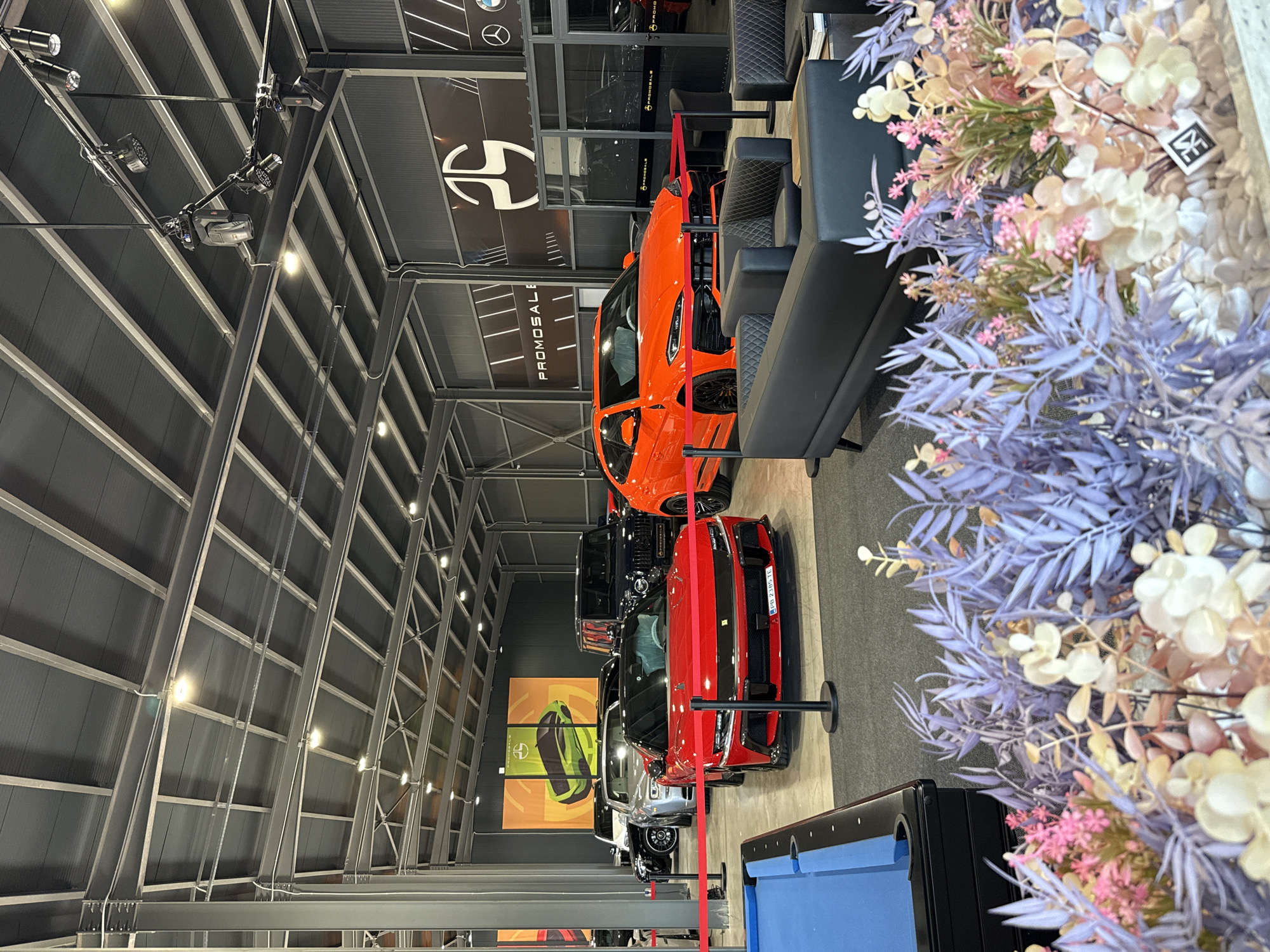 The interior of a dealership or showroom in Plovdiv, Bulgaria, is depicted, likely showcasing luxury vehicles. The building has an exposed steel beam structure. A line of cars, including an orange, red, black, silver, and white model, are parked in the center. Signs with the name "PROMOSALE" are visible. A black leather sofa and an artificial floral arrangement are at the side of the scene, and a blue pool table is in the foreground.