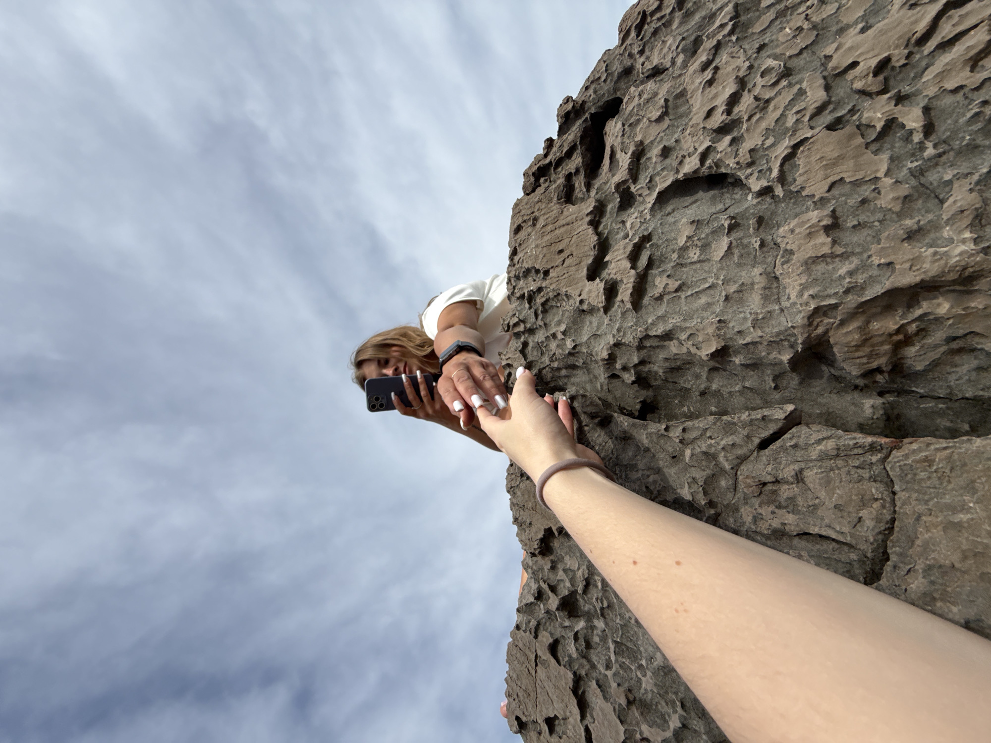A low-angle shot shows a person extending their arm and hand towards another person positioned at the edge of a rocky precipice. The person at the edge is holding a smartphone, seemingly taking a photo, and is reaching down with their other hand towards the extended hand. The location is identifiable as the city of Seoca in Montenegro, based on the unique geological features and rocky landscape. The sky with scattered clouds is visible in the background.