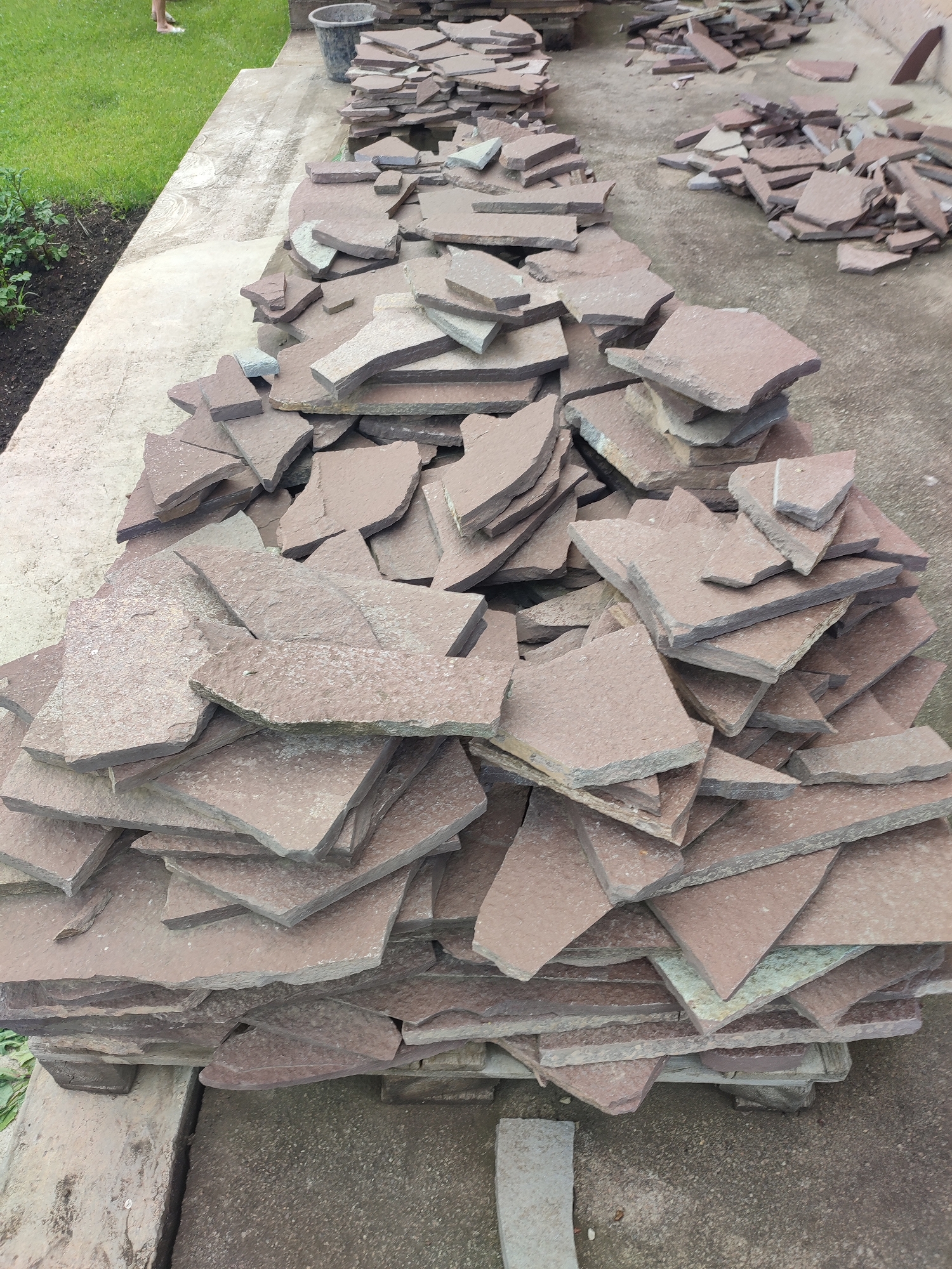 A collection of irregularly shaped, thin, reddish-brown stone slabs are piled on a concrete surface. The slabs vary in size and are stacked in multiple piles, with some laid out flat and others leaning against each other. The concrete surface is textured and appears to be an outdoor patio or walkway. To the left of the concrete, a patch of green grass and dark soil with some foliage is visible. In the background, a dark gray bucket and more piles of stone slabs can be seen. A blurred figure of a person with bare feet is visible standing on the grass in the upper left portion of the image. The overall scene depicts a construction or landscaping site where natural stone is being prepared or laid. This image was taken in Kumertau, Russia.