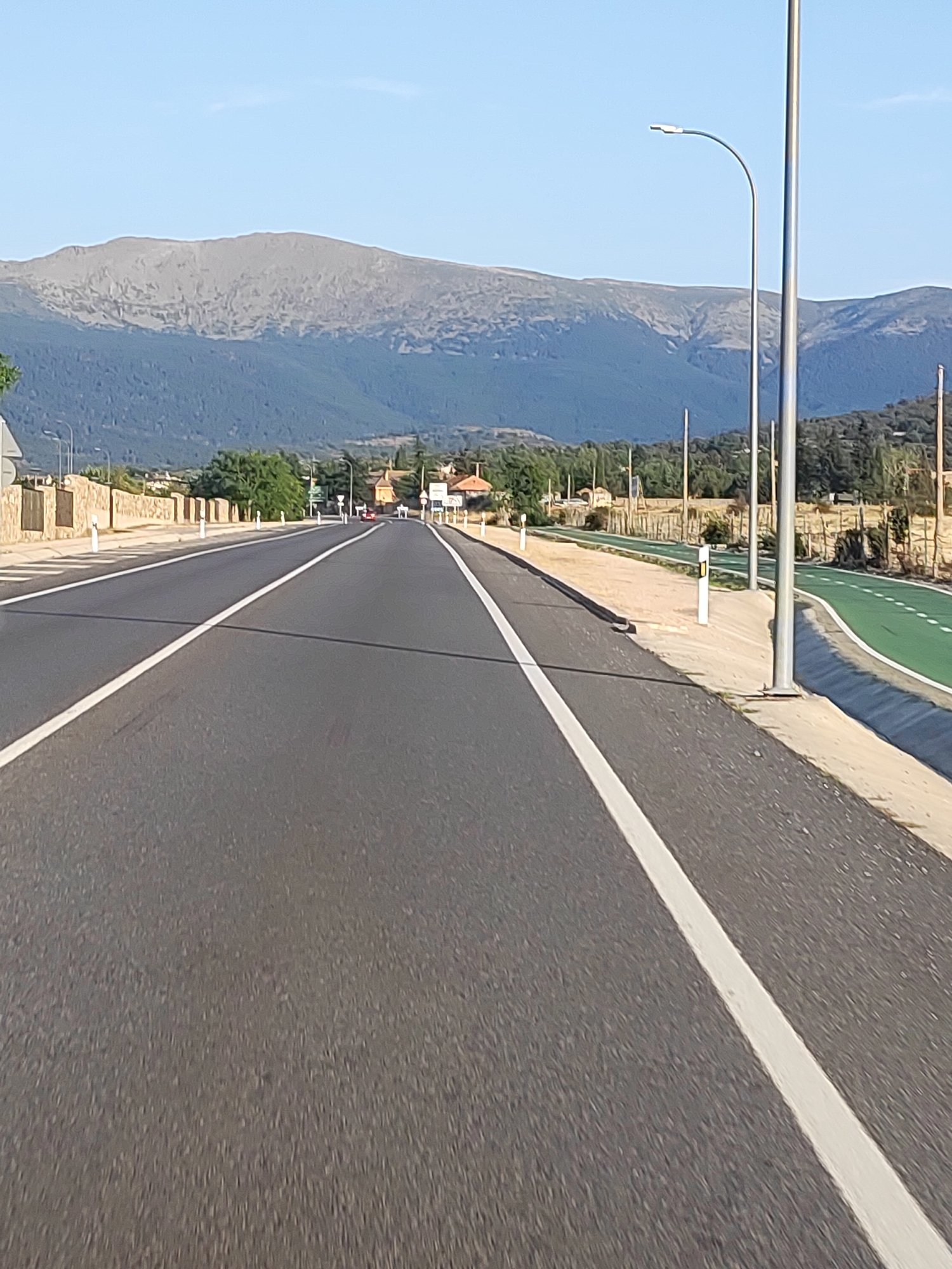 The image shows a straight, paved road with white lane markings leading towards a mountainous landscape under a clear blue sky. To the right of the road, there is a dedicated green lane, possibly for cycling, bordered by a sandy area with lampposts and reflective markers. A low stone wall runs along the left side of the road, behind which trees and some buildings are visible. The mountains in the background are covered in dark green vegetation at the base, transitioning to rocky, barren slopes at higher elevations. The lighting suggests it is daytime, likely in the morning or late afternoon, given the long shadows cast by the lampposts. There are no people or vehicles clearly visible in the immediate foreground or on the road itself.