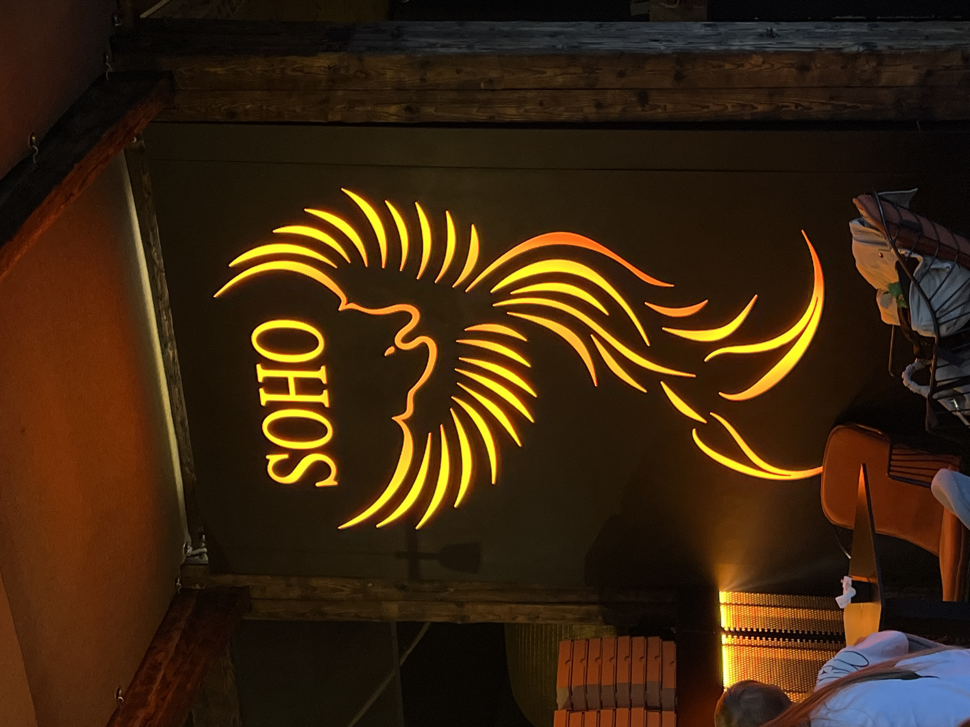 The image depicts a low-angle view of the "SOHO" sign in Bar, Montenegro. The sign is a dark backdrop with the word "SOHO" written vertically on the left in golden yellow letters. Next to the word "SOHO" is an illustration of what appears to be a lion's head or a stylized sun with curved rays extending outward, also in golden yellow. The sign is framed by dark wooden beams. In the lower right corner, there are elements of what seems to be a prop or display, possibly a stylized horse, and what seems to be a person wearing a white shirt looking up. The lighting is warm and bright, with the golden yellow of the sign contrasting sharply with the dark background. The location is Bar, Montenegro, based on the identifiable "SOHO" sign which is a local landmark.