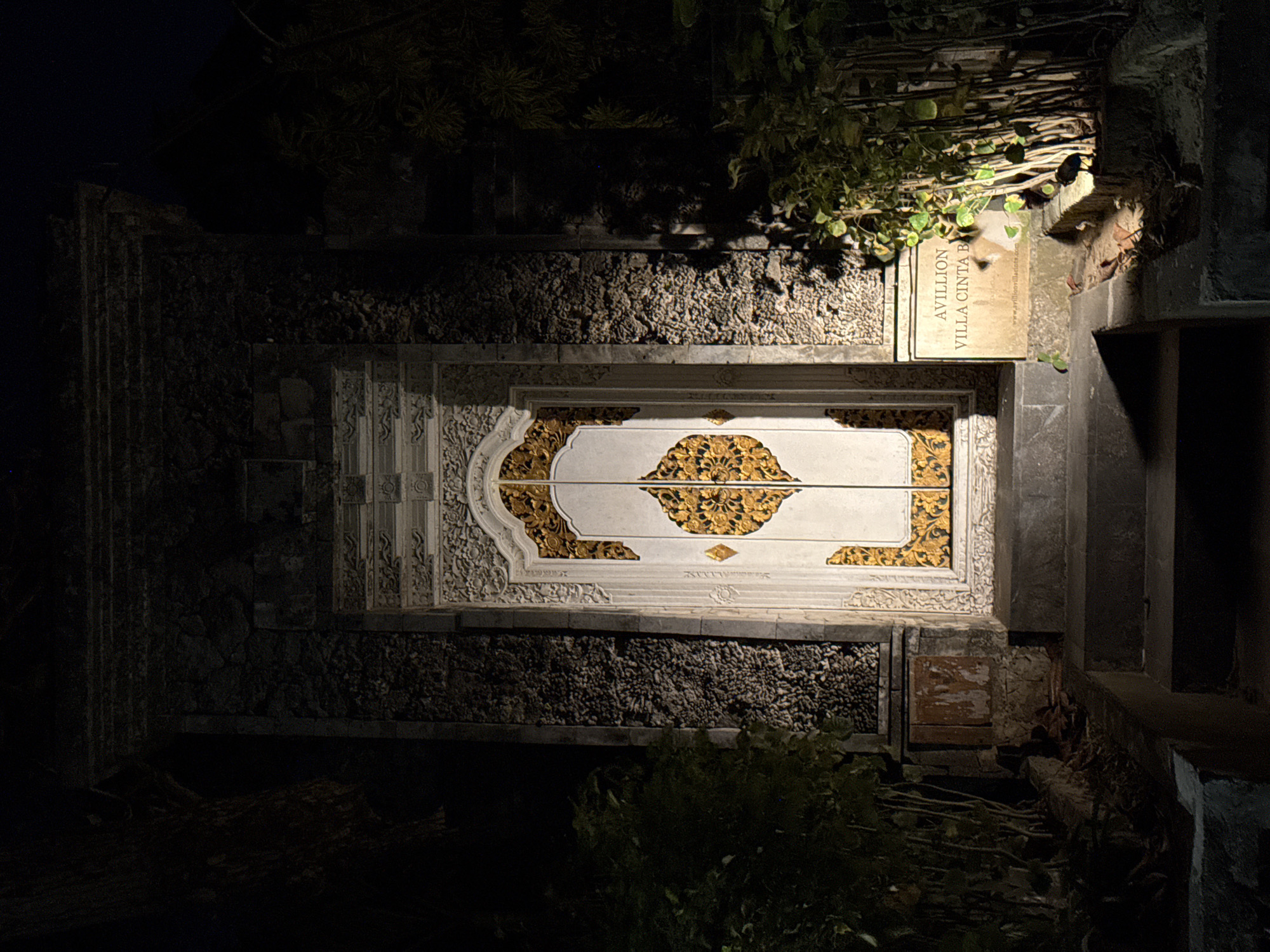 The image depicts the entrance to "AVILLION VILLA CINTA B" in Denpasar, Indonesia. A set of stone steps leads up to an ornate, white door with intricate gold carvings. The door is flanked by textured stone walls, and the area is illuminated by artificial lights, casting shadows and highlighting the architectural details. Lush green foliage surrounds the entrance. A sign to the right of the door displays the villa's name and website. The scene appears to be captured at night.