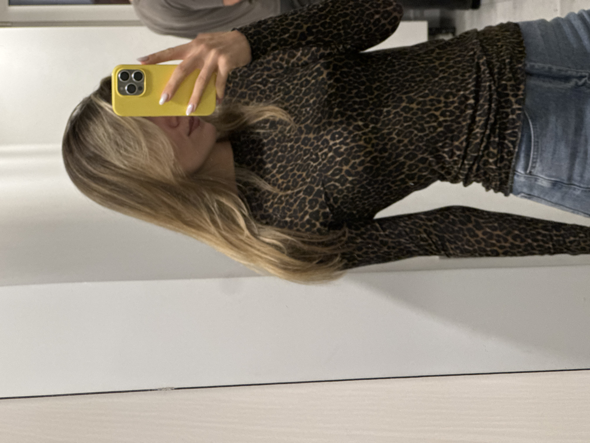 A young Caucasian woman with blonde hair, partially obscuring her face, takes a selfie in a mirror. She is wearing a long-sleeved, black and brown leopard print top and blue jeans. Her right hand holds a yellow smartphone in a yellow case. The background is a plain, light-colored wall. The scene appears to be indoors, possibly a fitting room or bathroom, in an unknown location.