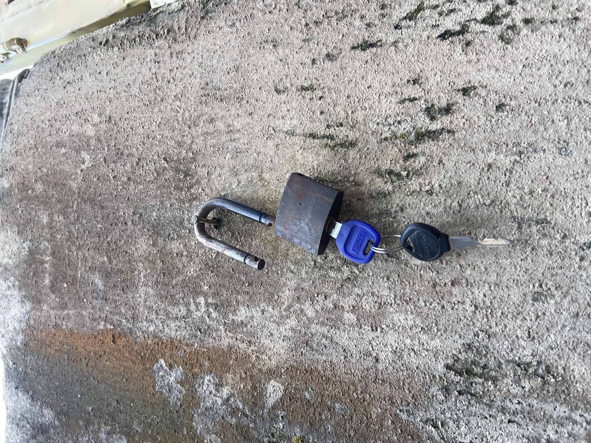 An unlocked, rusty metal padlock is attached to a rough, textured concrete wall in Mararaba, Nigeria. Two keys hang from the padlock's shackle. One key has a blue plastic head with the word "XFORCE" printed on it. The other key has a black plastic head with the word "haocure" printed on it. The concrete wall appears weathered, with some patches of discoloration and moss growth. A portion of a yellow structure is visible on the far right edge of the image.