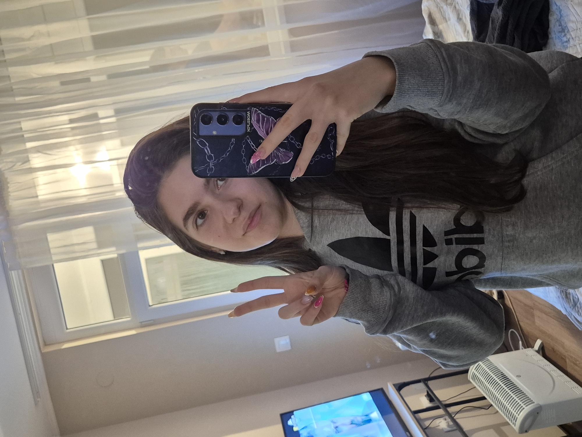 A young woman is taking a selfie in what appears to be a room in a residence. She is wearing a grey Adidas sweatshirt. She is holding a smartphone with a butterfly case in her right hand, making a peace sign with her left. The background includes a window with white curtains, a television, and a white electronic device. The location is Pavlikeni, Bulgaria.