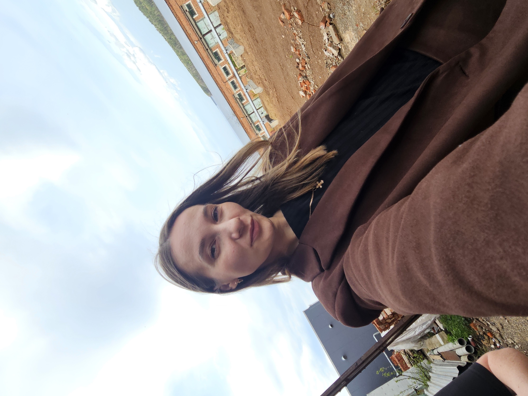 A woman is taking a selfie. She's wearing a brown coat over a black top. The location appears to be a rooftop or elevated construction site in Chaikovsky, Russia, as brick buildings and a body of water (possibly the Kama Reservoir) are visible in the background. There are construction materials like bricks, pipes, and corrugated metal sheets scattered around the site. The sky is partly cloudy.
