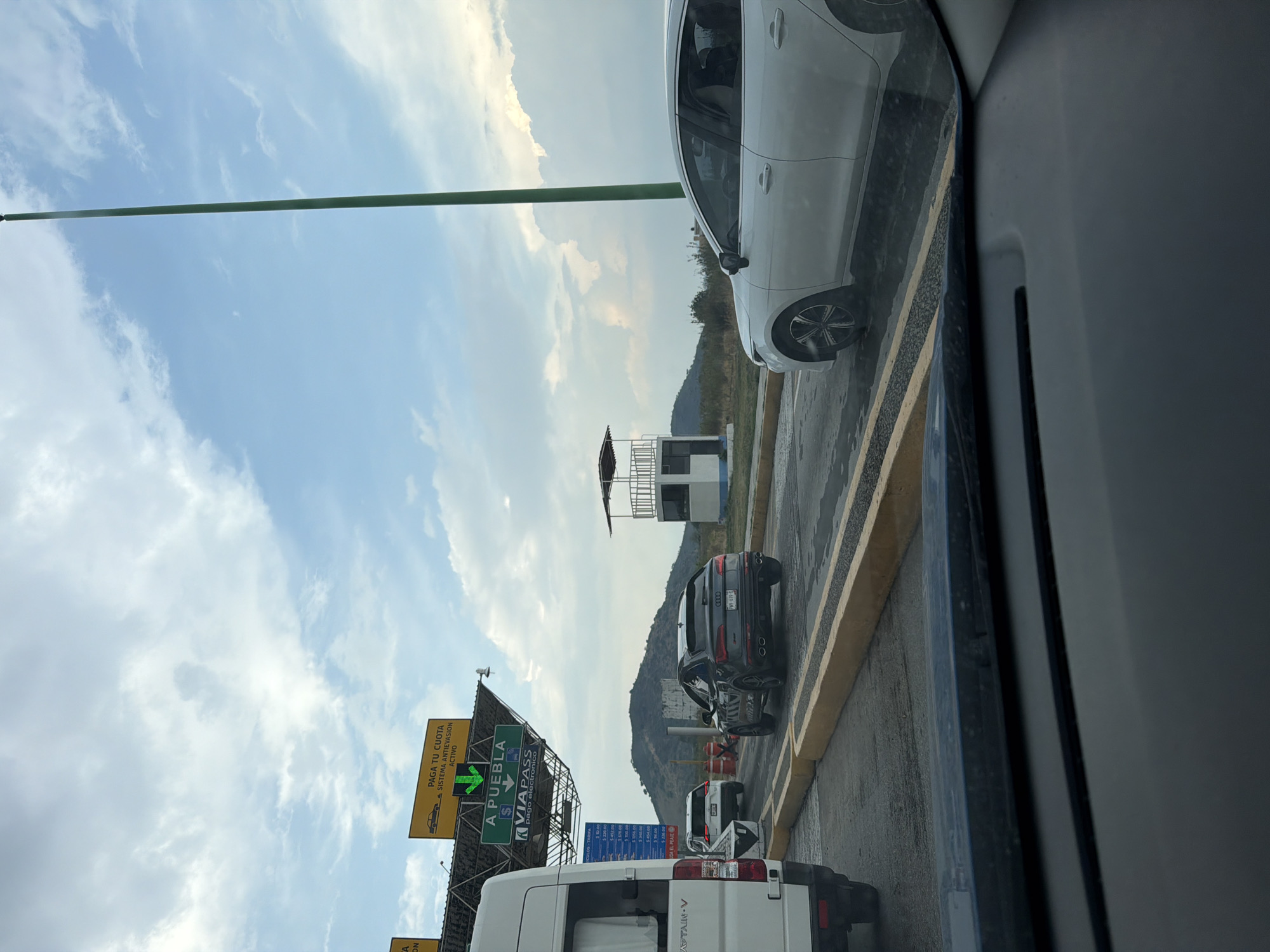 The image, taken from inside a car, shows a toll booth on a road in Metepec, Mexico. A white car is visible on the right, followed by an Audi SUV in the adjacent lane. A toll plaza structure is positioned between the vehicles. A sign indicating "Paga Tu Cuota" (Pay Your Fee) and direction towards Puebla is visible, along with the ViaPass electronic payment option. A white van is in the nearest lane. The sky is cloudy, and hills are visible in the background.