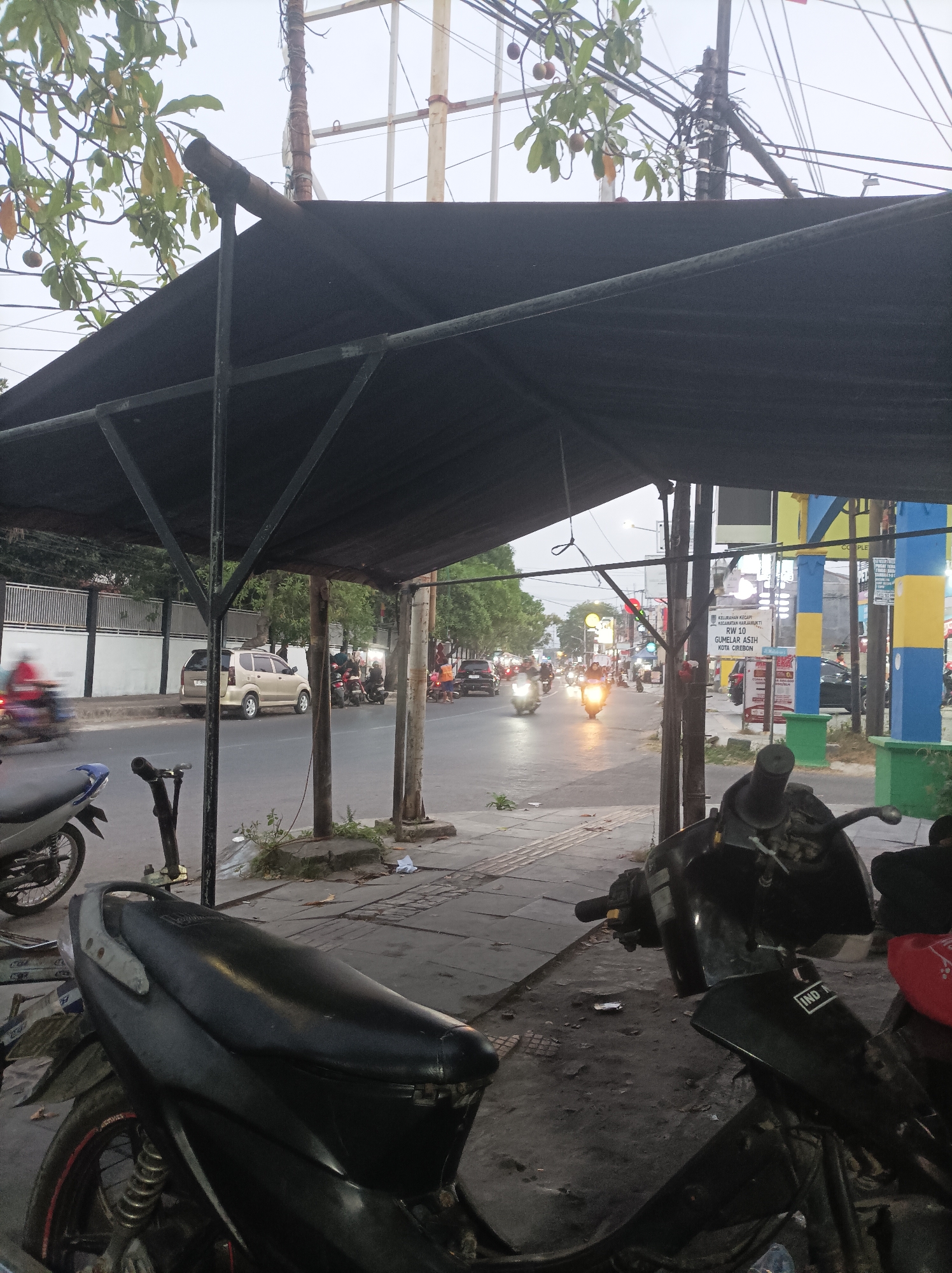 The image captures a street scene in Cirebon, Indonesia, likely during the late afternoon or early evening, as indicated by the dim lighting and streetlights beginning to illuminate. In the foreground, two motorcycles are parked under a dark canopy, which is supported by metal poles. The motorcycle on the left is partially visible, showing its rear wheel and fender. The motorcycle on the right is more prominent, with its black bodywork and handlebars clearly visible. A portion of its seat and a red object, possibly a bag or cushion, are also visible.

The street itself is paved, with a sidewalk running alongside it. Several trees with green leaves and some fruit are visible along the sidewalk. Beyond the sidewalk, the street is active with traffic. Cars and motorcycles are moving, with their headlights and taillights creating streaks of light, suggesting motion. Some of these vehicles appear to be stopped or moving slowly, possibly at an intersection.

In the background, there are various buildings and structures. One building on the left has a white wall with a dark fence. Further down the street, colorful structures, including pillars painted blue and yellow, are visible, suggesting commercial activity. Several signs are present, one of which, in the middle ground, is white with text in Indonesian, including "KELURAHAN ASAH", "RW 10", and "KOTA CIREBON", confirming the location as Cirebon. Overhead, there are numerous electrical wires and poles, a common sight in urban Indonesian settings. The sky is overcast. There are no clearly discernible individuals in sharp focus, though the movement of people within vehicles or on the sidewalk is implied by the general activity.