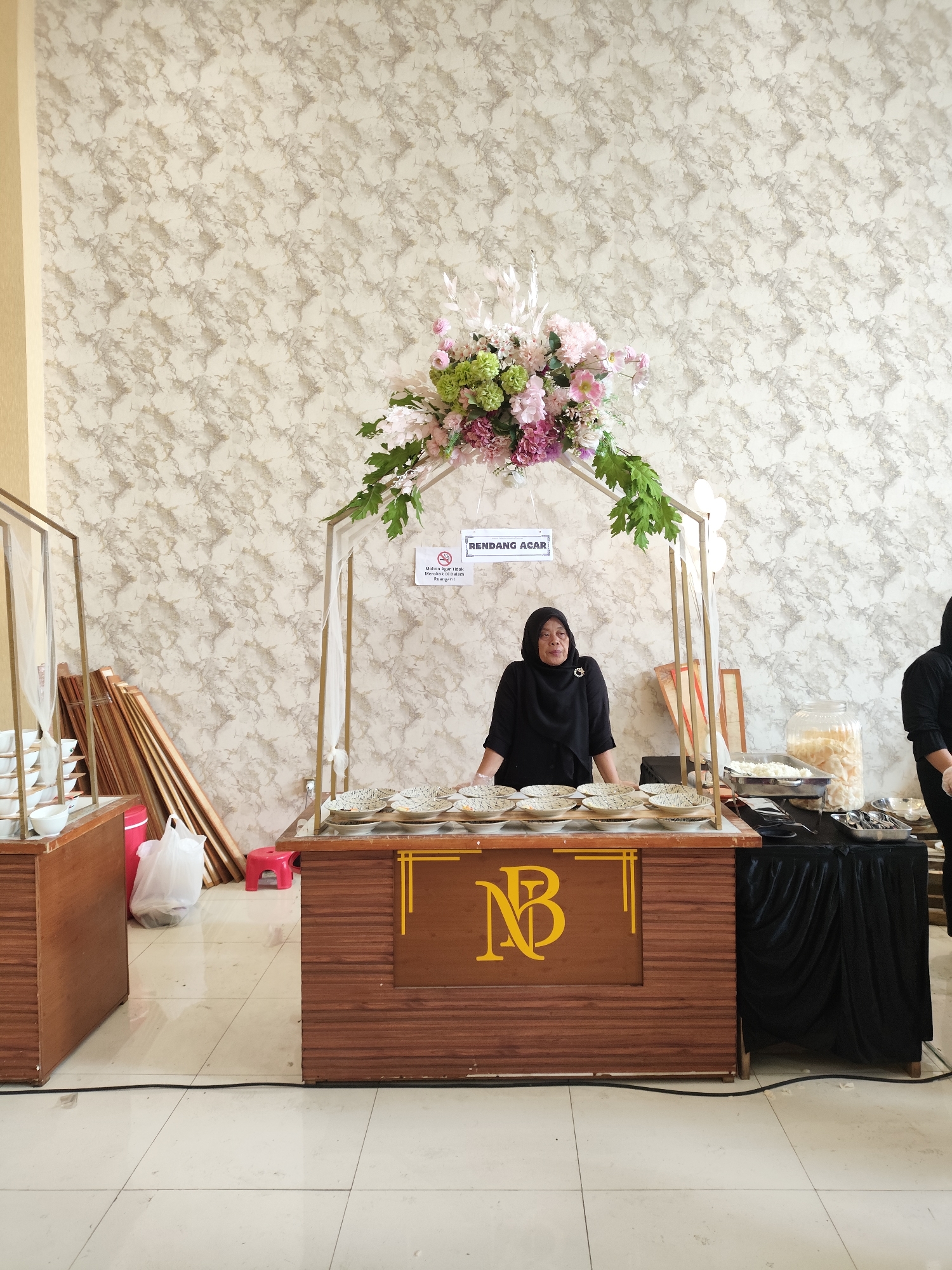 A woman wearing a black hijab and black clothing stands behind a wooden food stall in what appears to be an indoor venue. She is facing the camera and looking slightly to her left. The stall has a decorative metal arch above it, adorned with a large floral arrangement of pink, purple, and green flowers. A sign on the arch reads "RENDANG ACAR".
In front of the woman, several plates with patterned rims are stacked on the counter. To her right, a black tablecloth covers a surface, on which are food serving dishes, a large glass jar filled with white crackers, and cutlery. Another person, also wearing black, is partially visible to the far right, appearing to serve food.
The stall itself is made of dark wood with horizontal paneling. A yellow logo with the letters "NB" in a stylized font is visible on the front of the stall. To the left of the stall, another display unit with stacked white bowls is present. Various wooden planks and a white plastic bag are stacked on the floor behind the stall. The background features a textured wallpaper with a marble-like pattern in neutral tones. The floor is tiled with large, light-colored square tiles. The overall scene suggests a catering setup or a food service booth at an event. The location is identified as Dolopo, Indonesia.