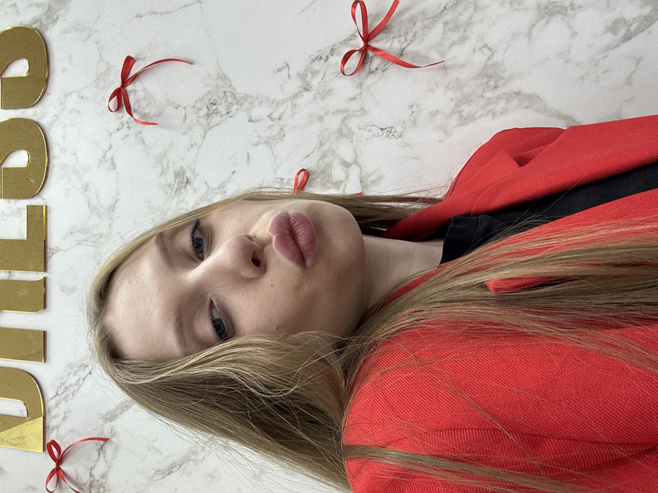 A blonde female individual is depicted in a close-up portrait, positioned slightly off-center to the left, within an indoor setting. She has blue eyes, fair skin, and is wearing red lipstick with her lips pursed in a pouting expression, directed towards the camera. Her long, light-colored hair falls over her shoulders. She is dressed in a red blazer worn over a black garment. The background consists of a white wall with a gray marble-like pattern. Partially visible at the top of the frame are gold-colored, reflective letters spelling "DRESS." Scattered across the wall are several small, red ribbon bows. The scene is consistent with the interior of a clothing retail establishment, such as a dress shop or boutique, in Chaikovsky, Russia.