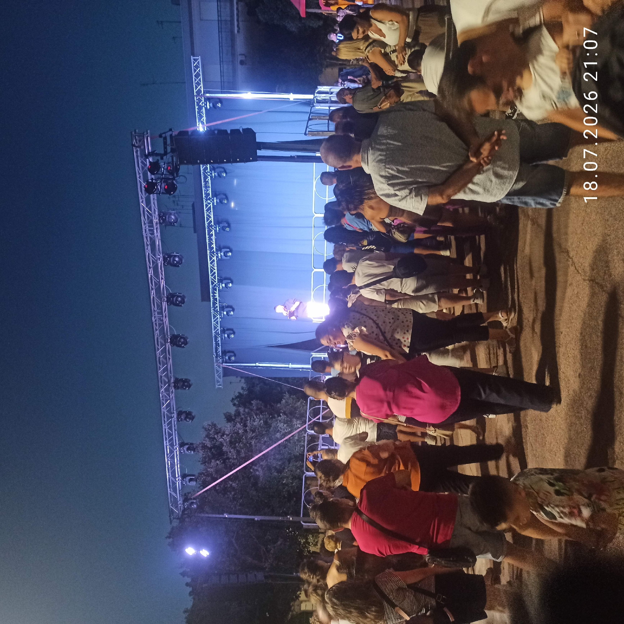 This is a medium-shot, eye-level, outdoor photograph taken at night, likely during a public event in the city of Bitonto, Italy. The foreground is filled with a crowd of people, viewed from behind and from the side, suggesting they are watching something. The main focus of the event is a stage in the background, illuminated by bright blue lights. A single performer, dressed in dark clothing, stands on the stage, holding a microphone and appearing to address the audience.
The stage is set up on a metal truss structure, with multiple spotlights mounted above. A large blue curtain forms the backdrop of the stage. In front of the stage, a metal barrier separates the performers from the audience. The ground appears to be paved.
The sky is dark, indicating it is night, with a faint gradient of blue. Trees are visible to the left and behind the stage. The overall atmosphere suggests a lively, open-air gathering. A date and time stamp, "18.07.2026 21:07", is visible in the lower right corner, though this may be a digital overlay and not reflective of the actual capture date.