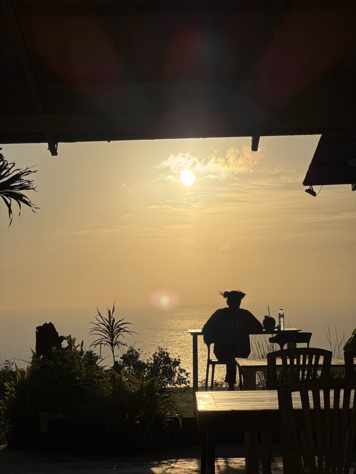 A silhouetted individual with hair tied in two buns is seated at a table overlooking the ocean at sunset. The person is wearing a shirt with the word "STAFF" visible. On the table, there is a drink in a coconut, a straw, and a bottle of water. The sun is setting over the horizon, casting a warm glow across the sky and water. Tropical foliage is visible in the foreground, and part of a wooden overhang is in the upper portion of the frame. The location is the city of Karon, Thailand.