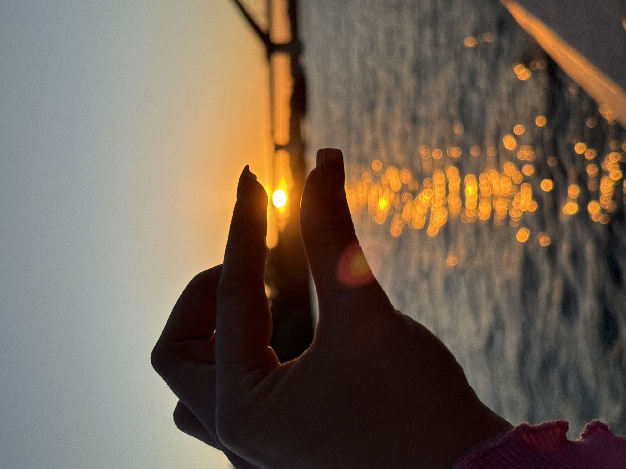 A hand is silhouetted against a sunset over a body of water, likely a river or lake. The sun is a bright orb peeking through the fingers. The water reflects the orange and yellow hues of the sunset, creating a shimmering effect. In the background, a bridge structure is visible. The image appears to be taken outdoors during golden hour. The location is not specified as Novosibirsk, Russia.