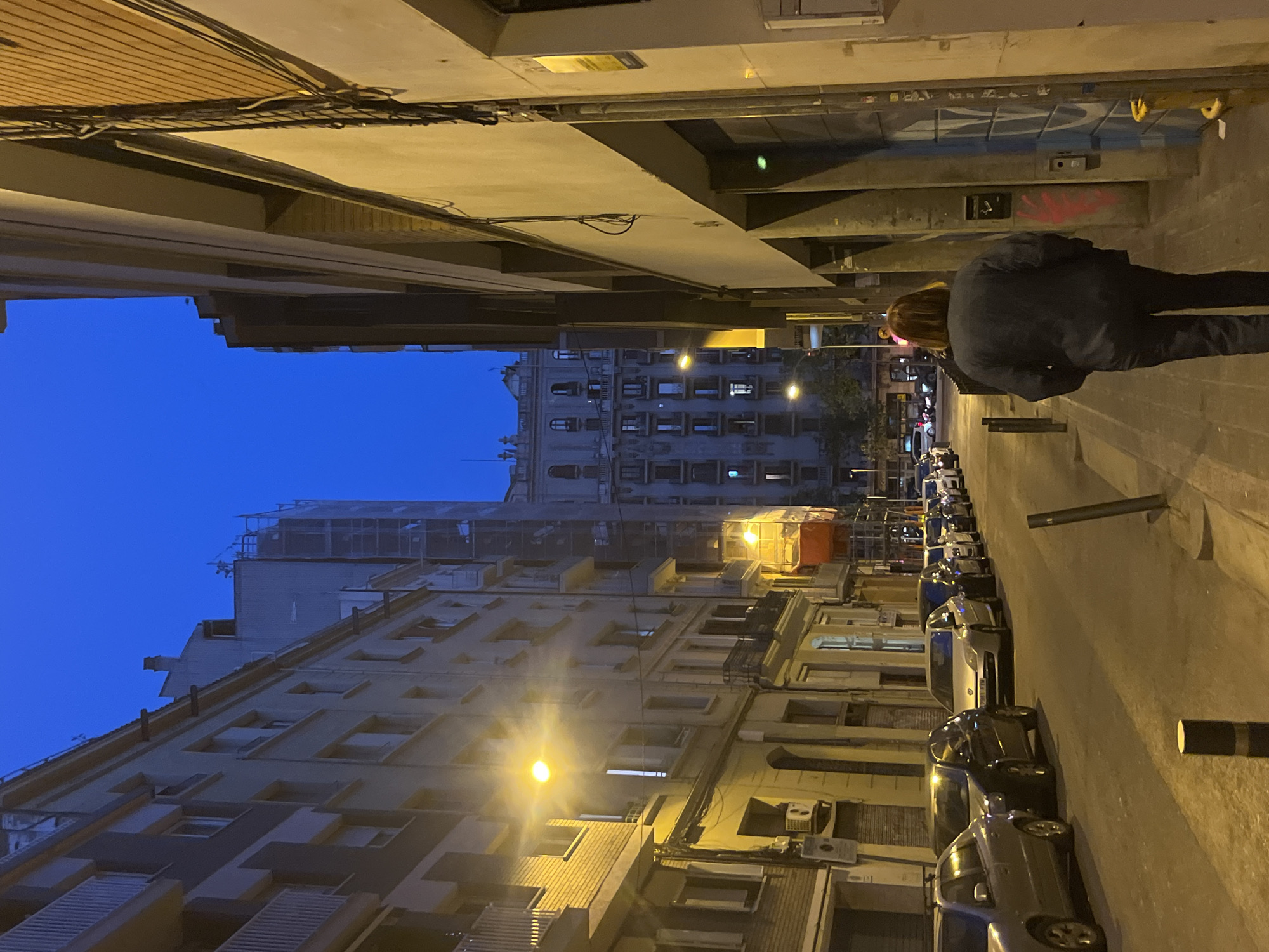 A nighttime urban scene, Barcelona, Spain, depicts a street with cars parked along the right side. Buildings of varying heights line the left side, illuminated by streetlights. One person, with shoulder-length brown hair, wearing dark clothing, stands on the sidewalk at the right. The perspective is angled upwards, capturing the sky above the buildings. A structure with scaffolding is visible atop one of the buildings.