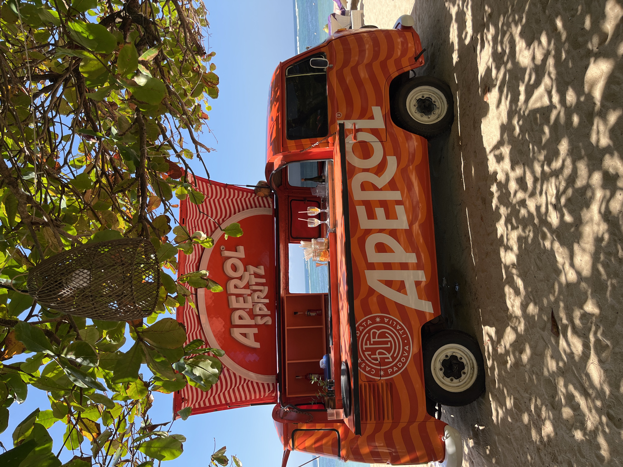 An orange Volkswagen Type 2 van, converted into a mobile Aperol Spritz bar, is parked on a sandy beach in Kuta Selatan, Indonesia. The van is decorated with wavy orange and red stripes and prominent "APEROL SPRITZ" branding. A red and white striped awning is extended above the serving window, where two Aperol Spritz cocktails with straws are visible. Behind the bar, bottles of Aperol and other bar supplies are arranged. The van has a circular logo on its side that reads "CASA DATA PADOVA ITALIA". The beach has light brown sand, and shadows from overhead tree leaves are cast across it. In the background, the ocean with gentle waves is visible, and a few beach chairs can be seen further down the shore. The sky is clear and blue. A woven light fixture hangs from a tree branch above the van.