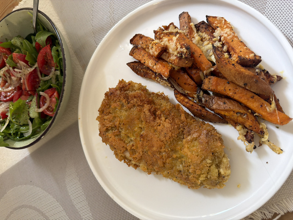 The image displays an overhead view of a prepared meal set on a light-colored, textured placemat, likely on a dining table. The specific location within Tel-Aviv, Israel, is not discernible from the visual information provided.
In the foreground, a white oval plate holds two main food items. On the left side of the plate is a large, golden-brown, breaded item, consistent with a fried chicken cutlet or schnitzel, exhibiting a visibly crispy texture. To the right of the cutlet, a pile of roasted sweet potato fries or wedges is present. These fries are orange-brown with some darker, charred edges, and are seasoned with dark specks, likely pepper or herbs, and topped with melted, light-colored cheese or similar granular garnish.
In the upper background, partially visible, is a light green ceramic bowl containing a fresh salad. The salad consists primarily of green leafy vegetables, sliced red tomatoes, and thin rings of white onion, appearing lightly dressed and seasoned. A metallic utensil, possibly a fork or spoon handle, is partially submerged in the salad. The overall environment suggests an indoor dining setting. No individuals are visible in the scene.