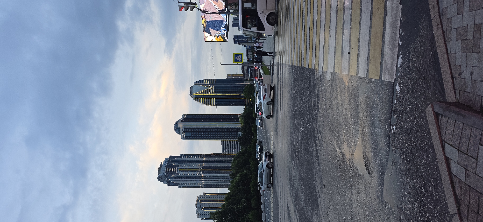 The image shows a street scene in Grozny, Russia. A row of modern high-rise buildings lines the left side of the street. These buildings feature architectural lighting and appear to be made of glass and steel. A row of green trees separates the buildings from the road. A street with multiple lanes for traffic and parked cars runs parallel to the buildings. A crosswalk with zebra stripes runs across the road. Pedestrians are seen walking on the crosswalk near a bus. Traffic signals and a billboard stand above the crosswalk. The sky is cloudy with hues of blue and gold near the horizon. A sidewalk made of paving stones occupies the right side of the image. The street appears to be wet, suggesting recent rainfall.