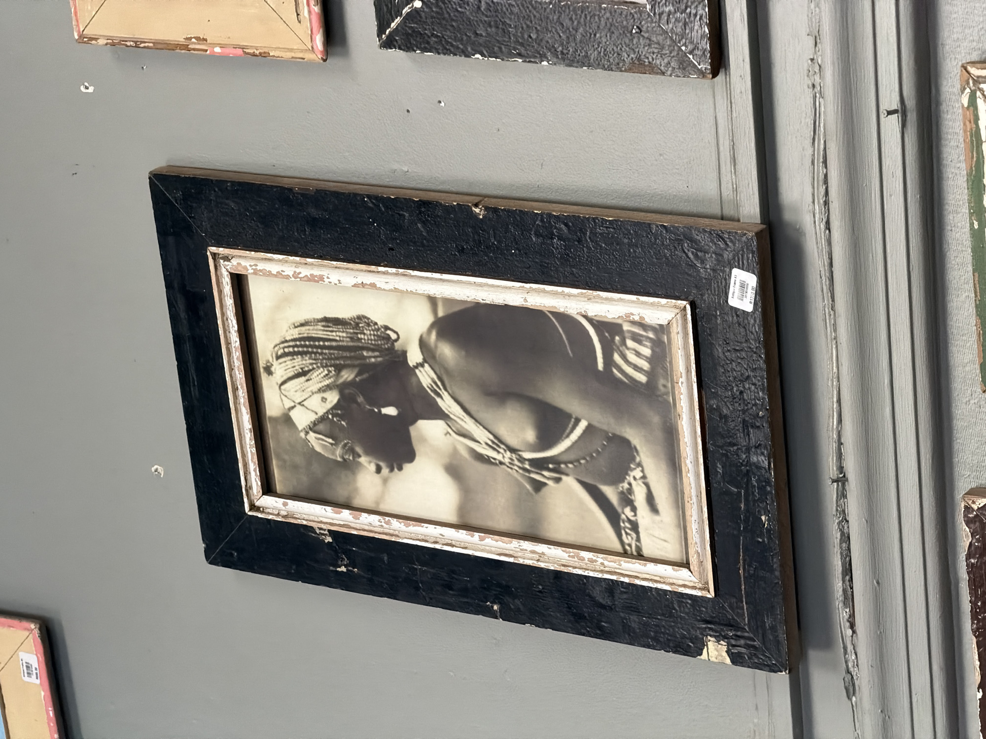 The central subject is a framed black and white photograph of a person, likely female, depicted in profile with their head bowed. The individual has intricately braided hair and is adorned with multiple strands of beaded necklaces or body jewelry. They appear to be in a focused pose, possibly engaged in an activity such as dancing or a ritual.
The photograph is housed in a rustic, chunky black frame with a heavily distressed and chipped finish, revealing lighter wood underneath. An inner white border, also distressed, shows hints of pinkish-brown. The frame is slightly tilted on the wall.
The scene is an indoor display, likely in an art shop or gallery, with framed items hung on a neutral gray wall. Other partially visible framed artworks surround the main piece, including a light pink/beige frame above, another similar dark frame, and a green/brown distressed frame to the right. To the far right, there is a section of vertical wooden paneling, painted a lighter gray and exhibiting signs of wear and peeling paint, with a nail partly embedded in it. The overall aesthetic of the frames and wall suggests a vintage or rustic style.
The black and white photograph features a softly blurred background, drawing focus to the figure. Small imperfections and tiny holes are visible on the gray wall. A white sticker with a barcode is affixed to the upper right corner of the main frame, displaying the price "R 250.00", indicating South African Rand.