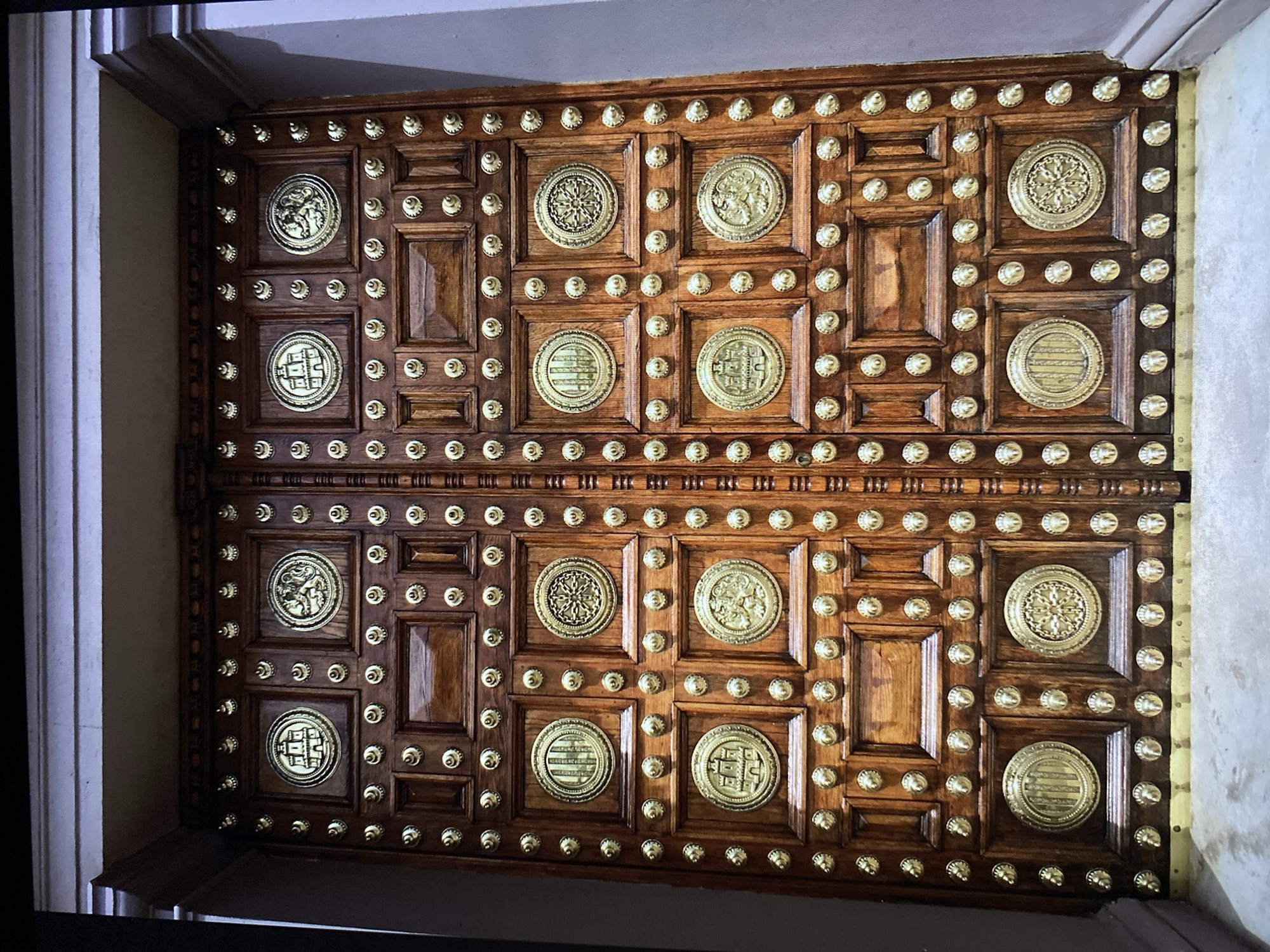 A large, ornate, wooden double door is shown in full view. The doors are constructed from rich, dark wood and are heavily decorated with numerous brass studs and circular brass medallions. The medallions are intricately designed with various patterns, including what appear to be architectural structures, heraldic symbols such as lions and eagles, and geometric patterns. The doors are divided into multiple rectangular panels, each containing these decorative elements. The top of the doors are framed by a dark wooden architrave, which sits against a lighter colored wall. The floor in front of the doors appears to be concrete or stone, with a brass kick plate at the bottom of the doors. The lighting suggests an indoor setting, possibly a grand entrance to a historical building. The image captures the intricate craftsmanship and historical significance of the doors.