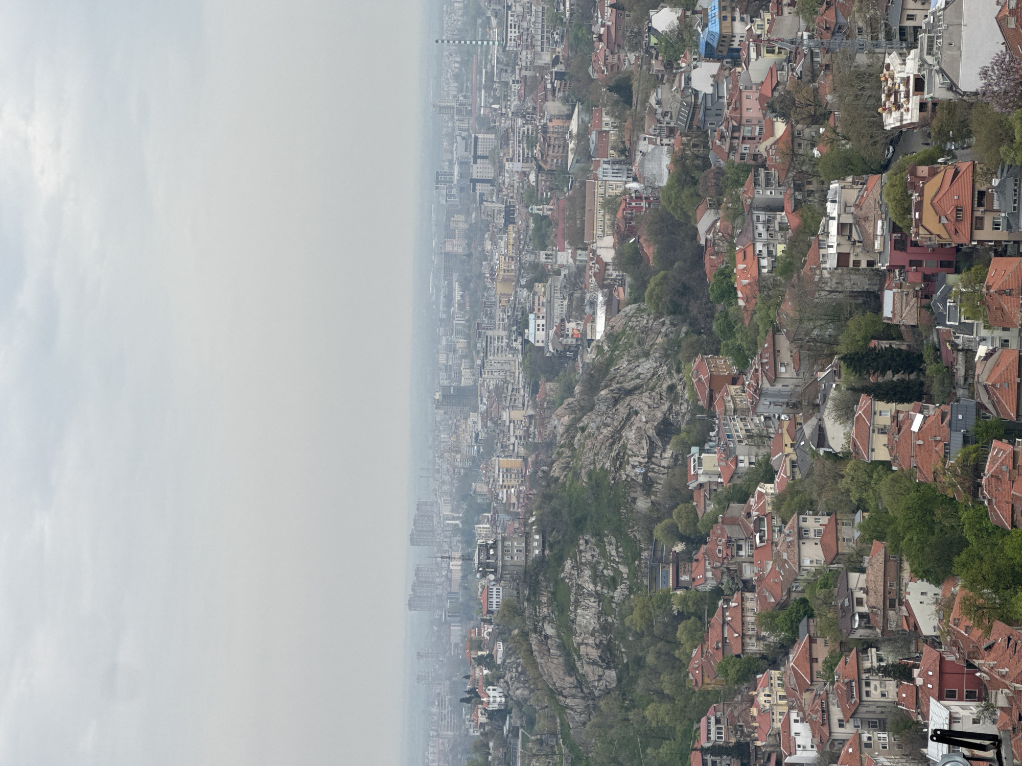 This aerial or high-vantage-point image captures a sprawling cityscape, identified as Plovdiv, Bulgaria, under an overcast and hazy sky. The photograph is oriented vertically, showing the horizon line running diagonally across the frame, with the sky dominating the upper left and the city stretching across the lower right.
The scene is characterized by a dense urban environment built around and upon prominent, rocky hills. These natural formations, covered with sparse green vegetation and bare trees, divide and integrate with the residential areas. The city itself presents a mosaic of architectural styles, predominantly featuring buildings with traditional red-tiled gabled roofs, particularly on the hillsides and in the foreground. These are interspersed with more modern, multi-story buildings and apartment blocks that become increasingly dense and faded by the atmospheric haze towards the distant horizon.
Numerous trees and green spaces are visible throughout, especially clinging to the rocky terrain and dotting the residential areas. In the middle ground, a tall, striped industrial chimney stands out against the muted backdrop of the city. Roads and infrastructure weave through the urban fabric, though specific details are obscured by distance and atmospheric conditions. A construction crane can be faintly observed in one of the more detailed sections.
The time of day appears to be daytime, but the heavy overcast sky creates a soft, diffused light without clear shadows. The weather is hazy or misty, reducing visibility and giving the distant cityscape a desaturated, uniform grey tone. There are no people or specific activities discernible in this broad overview. No visible text is present in the image.