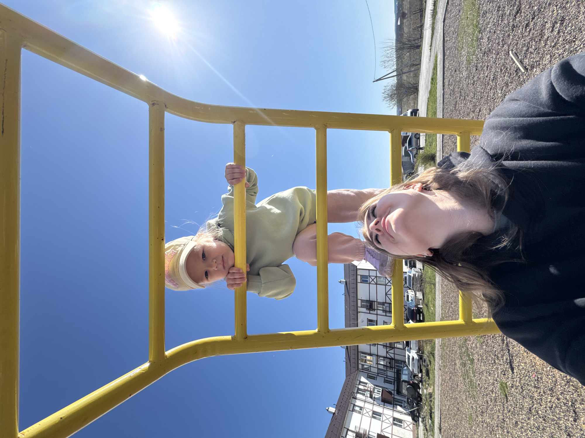 A low-angle shot taken in Горный, Russia, shows a young child hanging upside down on a yellow metal jungle gym, while an adult woman is standing nearby and appears to be taking a selfie. The child is wearing a light green sweater and a light-colored hat. The woman is wearing a dark hoodie and has brown hair. The background shows a clear blue sky, a row of white and brown buildings, a gravel area, a road, and some trees. The sun is shining brightly.