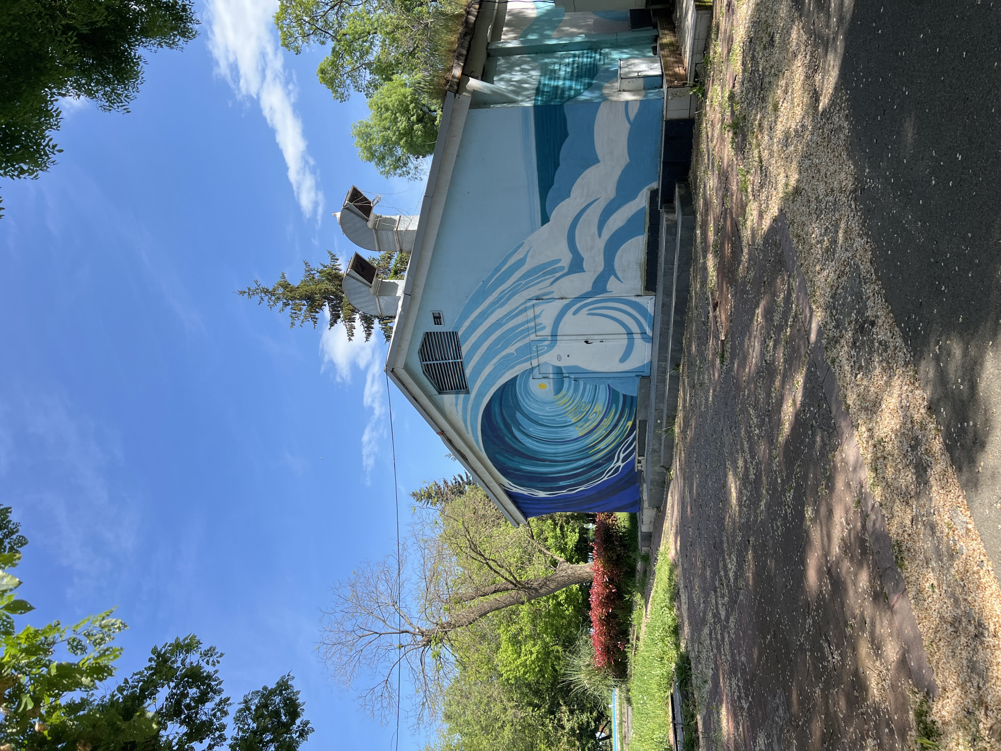 A view of a light blue building with a painted ocean wave mural on the side, likely taken outdoors in a public space. The building has two visible vents on the roof. A brick or paved pathway runs alongside the building, and there is greenery, including trees and bushes, in the foreground. The sky above is blue with scattered clouds. The location is Burgas, Bulgaria.