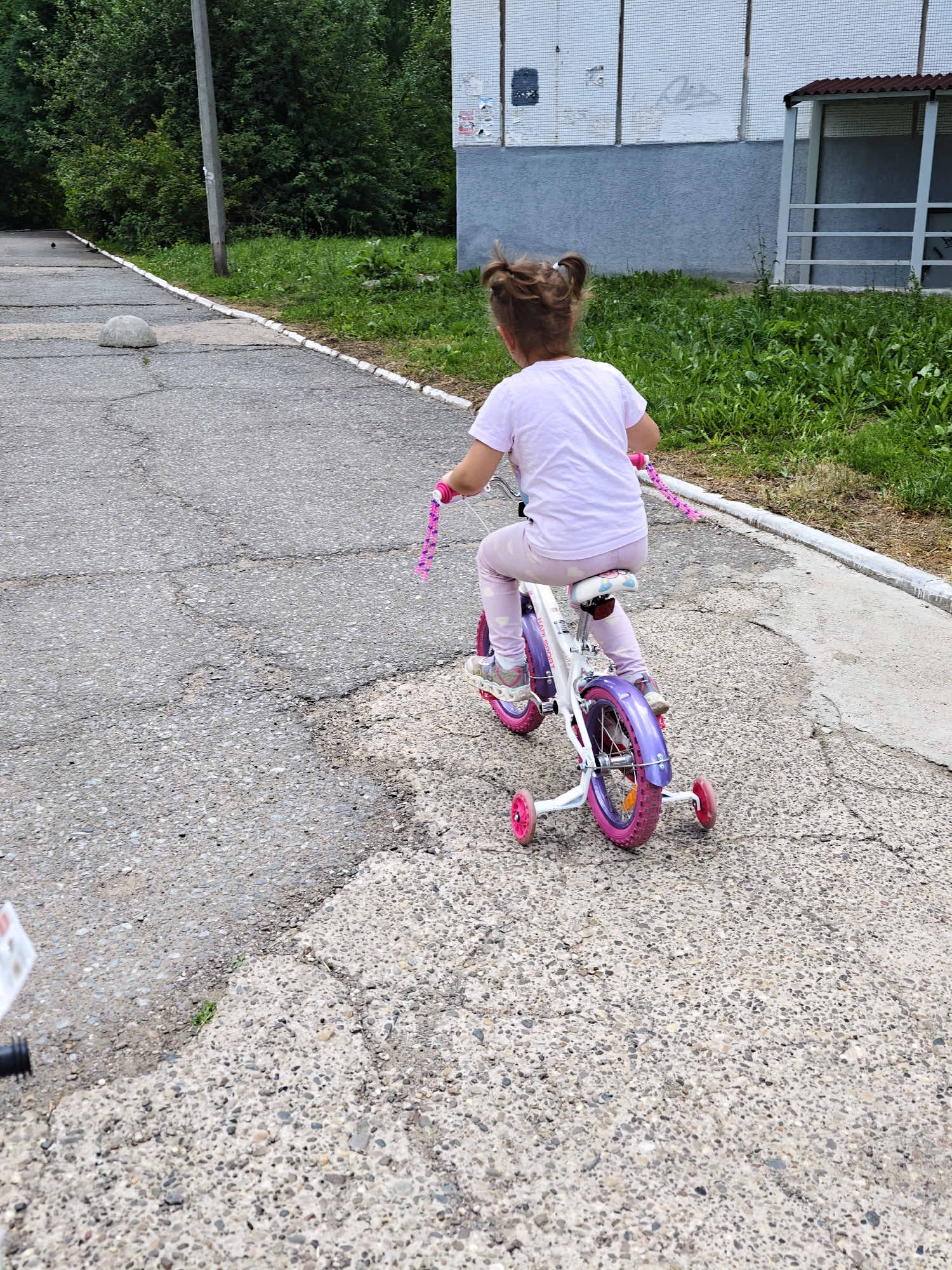 A young girl, seen from behind, rides a white and purple bicycle with pink accents and training wheels on a cracked asphalt path. She wears a light pink t-shirt and light pink pants, with her brown hair styled in pigtails. The path is bordered by grass and a white curb on the right side. In the background, there is a building with light-colored walls and a brown roofed structure. A utility pole stands on the left side of the path, and trees are visible behind it. The scene appears to be in a residential area, possibly a courtyard or park in Chaikovsky, Russia.