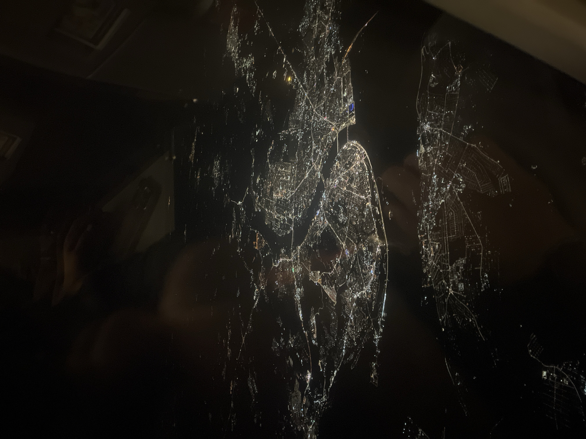The image depicts a night-time view of city lights, possibly Морозовка, Russia, as seen through a reflective or refractive surface, creating a distorted or broken effect. The city lights appear as scattered points and lines of light against a dark background. The reflection or refraction causes the lights to appear fragmented and dispersed, as if viewed through a broken mirror or water surface. The foreground is dark and may show reflections of objects or people.