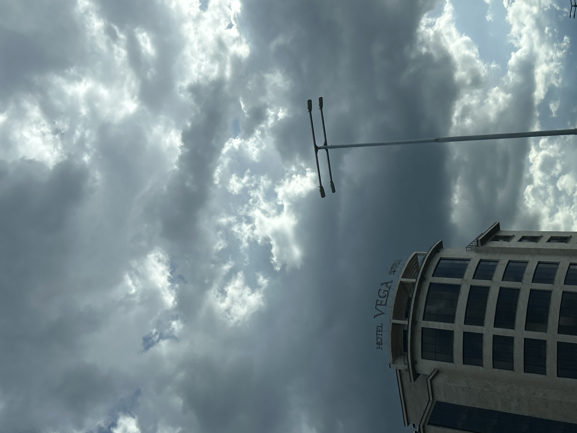 The image shows an upward-angled view of the Hotel Vega Sofia in Sofia, Bulgaria, against a cloudy sky. A tall, gray light post is visible near the top of the frame, slightly to the right of the building. The hotel's facade is modern, with multiple rows of windows. The sky is overcast with a mix of light and dark clouds. The image appears to have been taken from a low vantage point, looking up at the building and the sky.