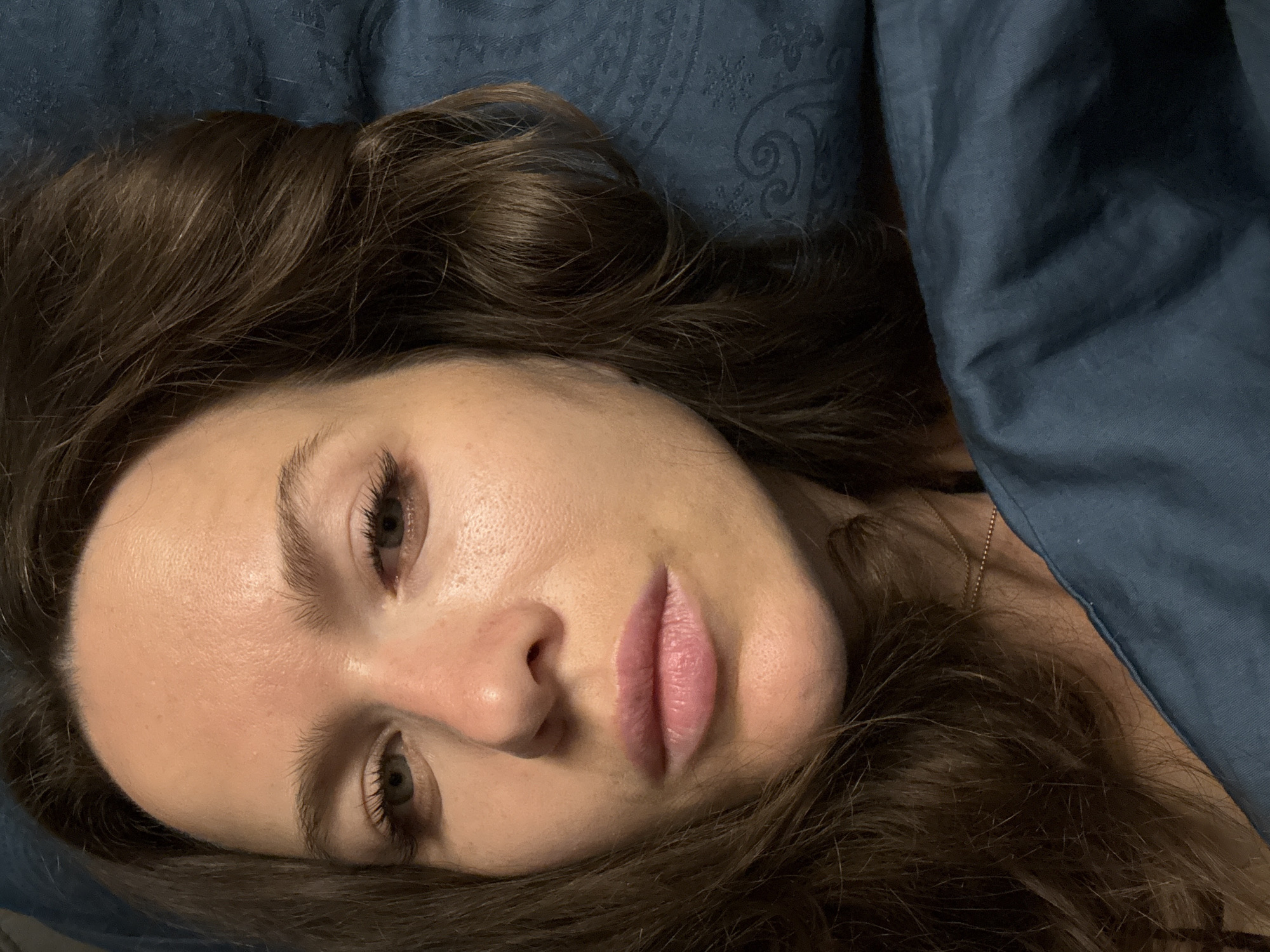 A close-up, portrait-style photograph depicts a woman lying on a blue bed or pillow. Her head is tilted to the right, with her dark brown hair spread around her. She has fair skin, defined eyebrows, and full lips. Her eyes are partially open, and she appears to be looking at the camera. A blue blanket or sheet is draped over part of her shoulder and upper body. The location of the image is not evident.