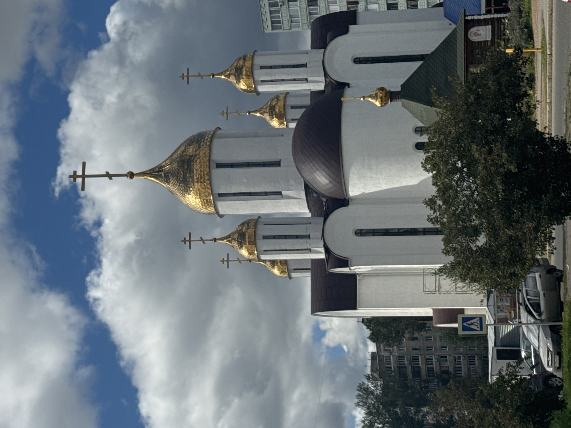The image shows the Ascension Cathedral in Ufa, Russia. The cathedral is a large, white building with multiple golden domes topped with crosses. In front of the cathedral is a smaller, wooden chapel with a green roof and a golden dome. There are trees and bushes in the foreground, as well as cars parked on the side of the road. In the background, there are apartment buildings. The sky is blue with white clouds.