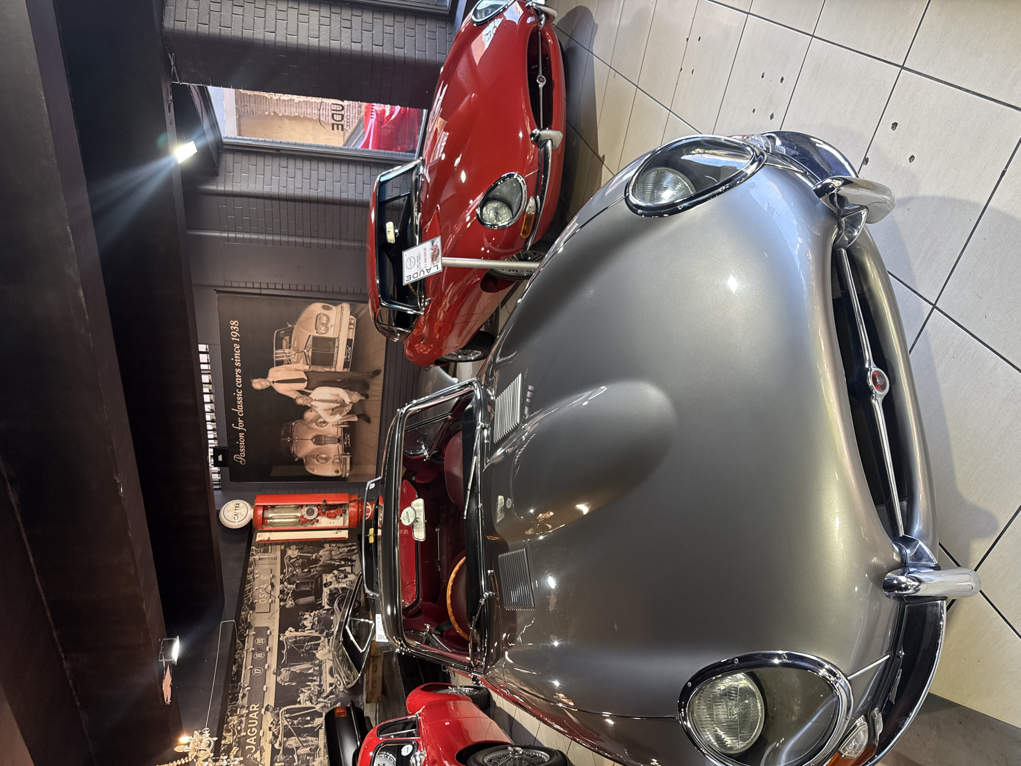 This image captures an indoor display of classic cars, likely within a showroom or museum in Cape Town, South Africa. The setting features a light-tiled floor and dark, possibly black, walls adorned with large historical photographs and text. Bright overhead lights illuminate the space, creating prominent reflections on the polished car surfaces.
The primary subjects are two classic Jaguar E-Type cars. In the foreground, positioned to the right, is a silver or light grey convertible model, highly polished and reflective. Its sleek lines, chrome bumper, headlight surrounds, and side mirrors are clearly visible. The interior, partially seen, features red leather upholstery and a wooden steering wheel. Behind and to the left of the silver car is a bright red Jaguar E-Type, appearing to be a coupe, with its front-left section partially obscured by a white sign on a stand. Another red car is briefly visible at the bottom of the frame.
The dark walls serve as a backdrop, featuring large black and white photographs depicting older classic cars and historical figures, possibly mechanics or enthusiasts. One large poster reads, "Passion for classic cars since 1938." Another section of the wall includes the word "JAGUAR" and a timeline-like graphic with numbers "17," "18," "19," and "20." A vintage red "CALTEX" fuel pump adds to the nostalgic automotive theme. A small white circular clock is also mounted on the wall. Text on a sign near the red car, and also on a wall poster, indicates "LAUDE." No live people are visible in the immediate scene.
The image presents a static exhibition, emphasizing the aesthetic and historical significance of these iconic vehicles in a dedicated, well-lit environment.