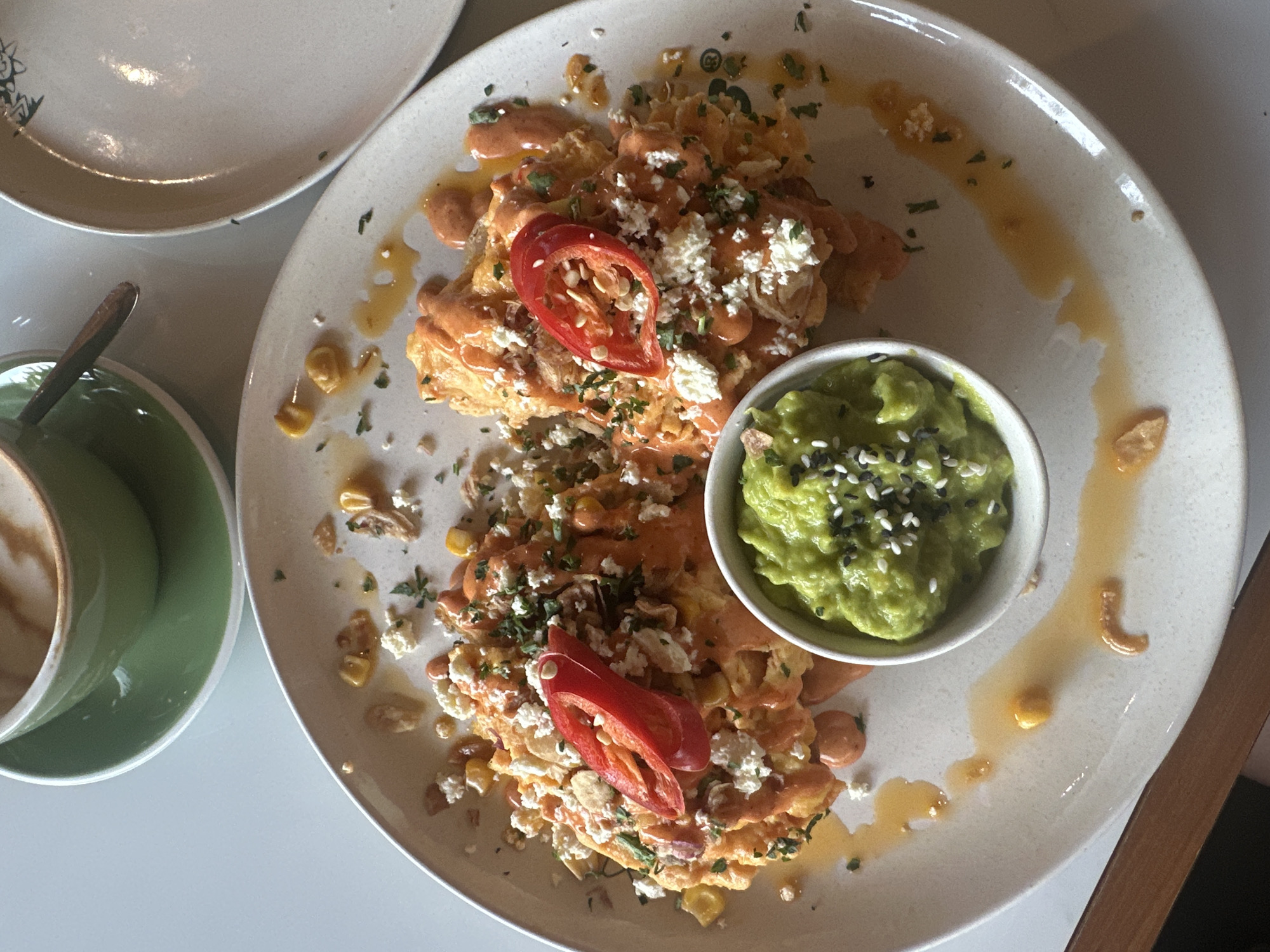 A top-down, close-up shot shows a partially eaten breakfast plate next to a cup of coffee. The plate contains what appears to be scrambled eggs with sauce, crumbled cheese, corn, parsley, and two slices of red pepper, with a separate small bowl of guacamole topped with sesame seeds. The food is presented with sauce drizzled artistically on the side of the plate. Next to the plate is a green cup of coffee with a silver spoon. Another, empty plate is visible in the upper left of the image. The background is blurred, suggesting a shallow depth of field. The location is Mengwi, Indonesia.
