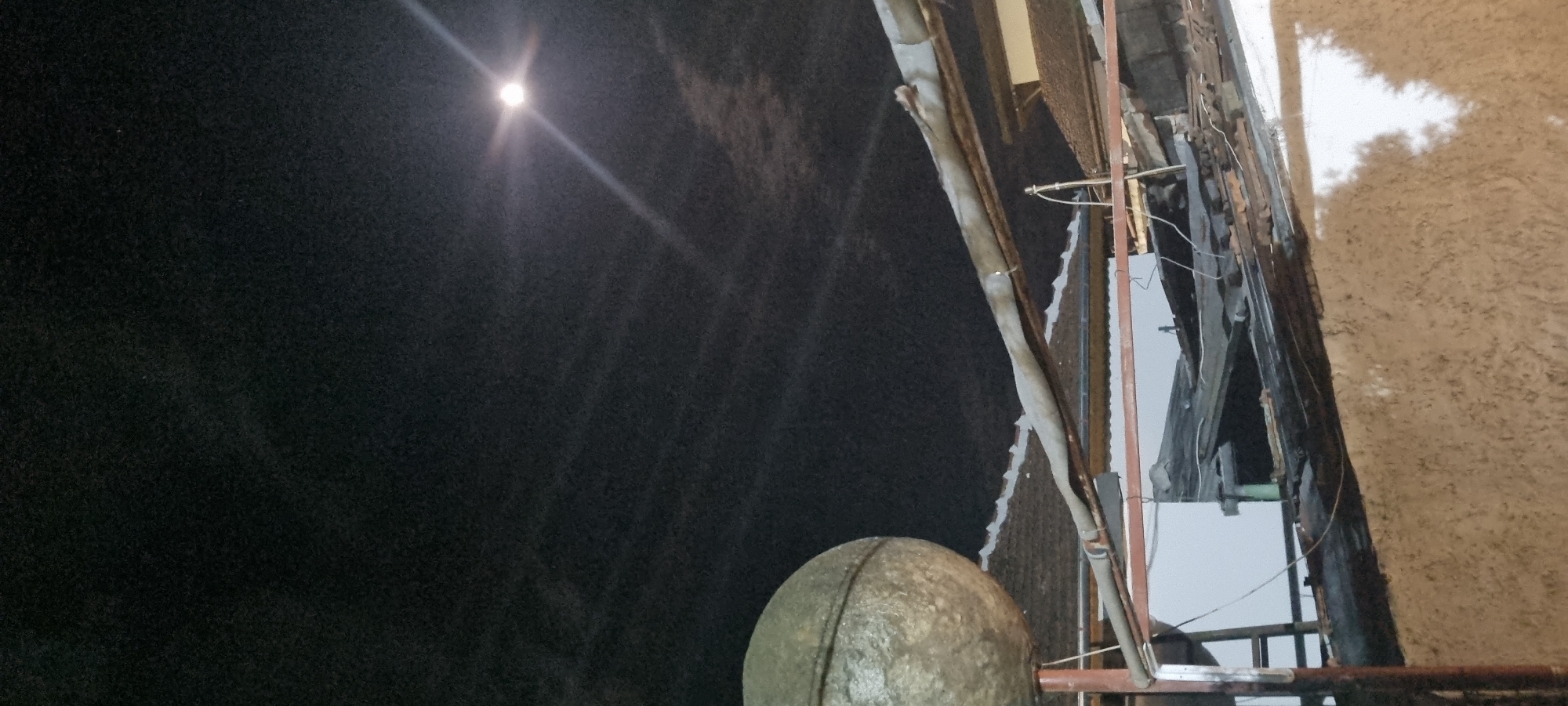 The image depicts an exterior night scene under a bright light source resembling the moon. A large, spherical object dominates the lower center, possibly a concrete structure or a weathered sphere-shaped decoration. To the right of the sphere, there is a complex arrangement of pipes, wires, and what appears to be construction scaffolding, including metal beams and weathered wooden elements, suggesting ongoing repairs or demolition. The right side of the scene features a rough, textured wall or surface, possibly made of stucco or a similar material. The left side is characterized by deep shadows and minimal details. The location in the city of Unknown, Unknown is indeterminate from the provided image.
