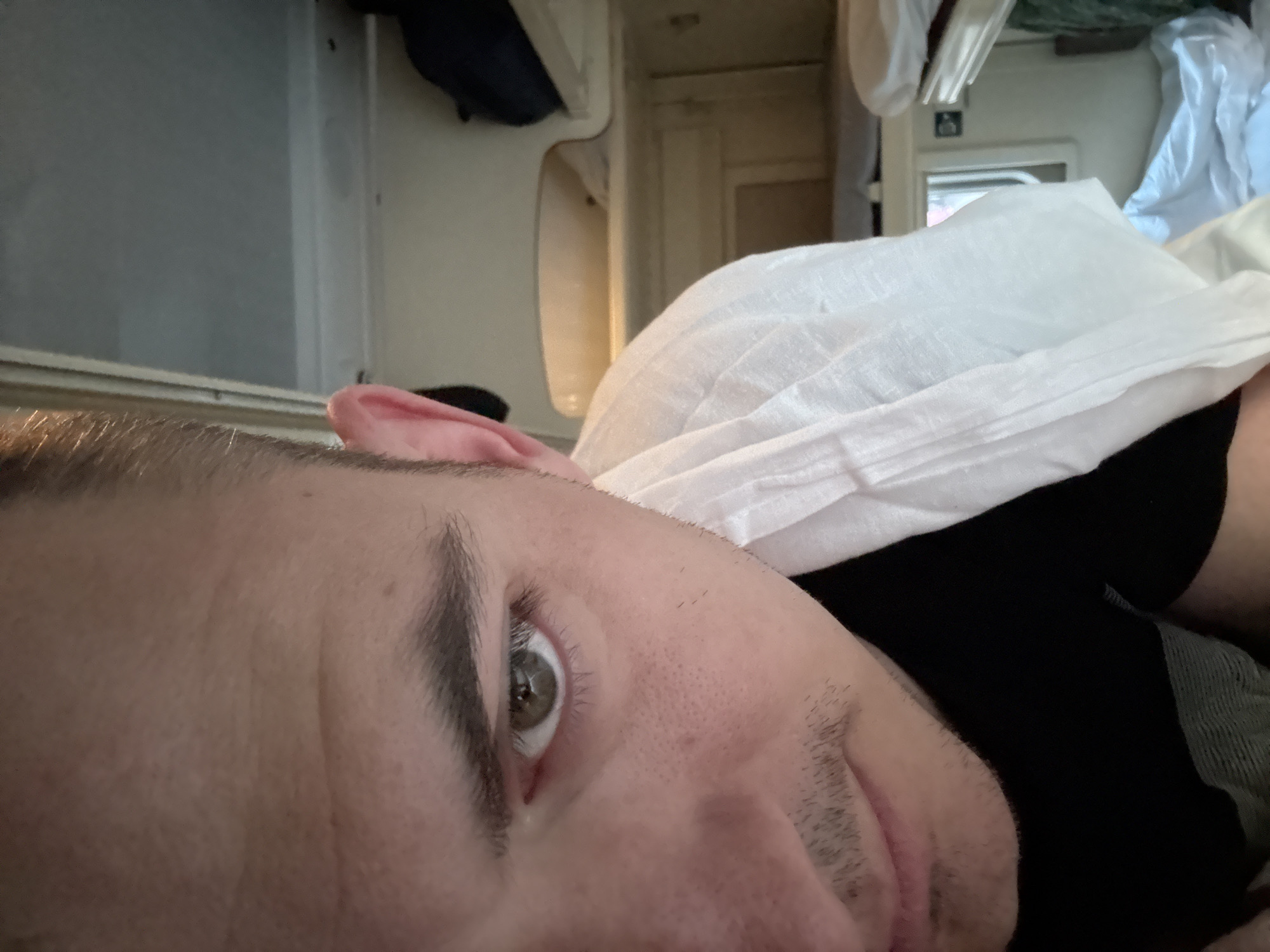 A close-up, angled perspective captures a person lying down, possibly on a bed, with their face turned towards the viewer. A white pillow partially covers the person. The surrounding environment appears to be indoors, with a view of what could be the interior of a recreational vehicle or a small living space. Given the level of detail visible in the image, the city of Ryazan, Russia cannot be determined from the picture alone.