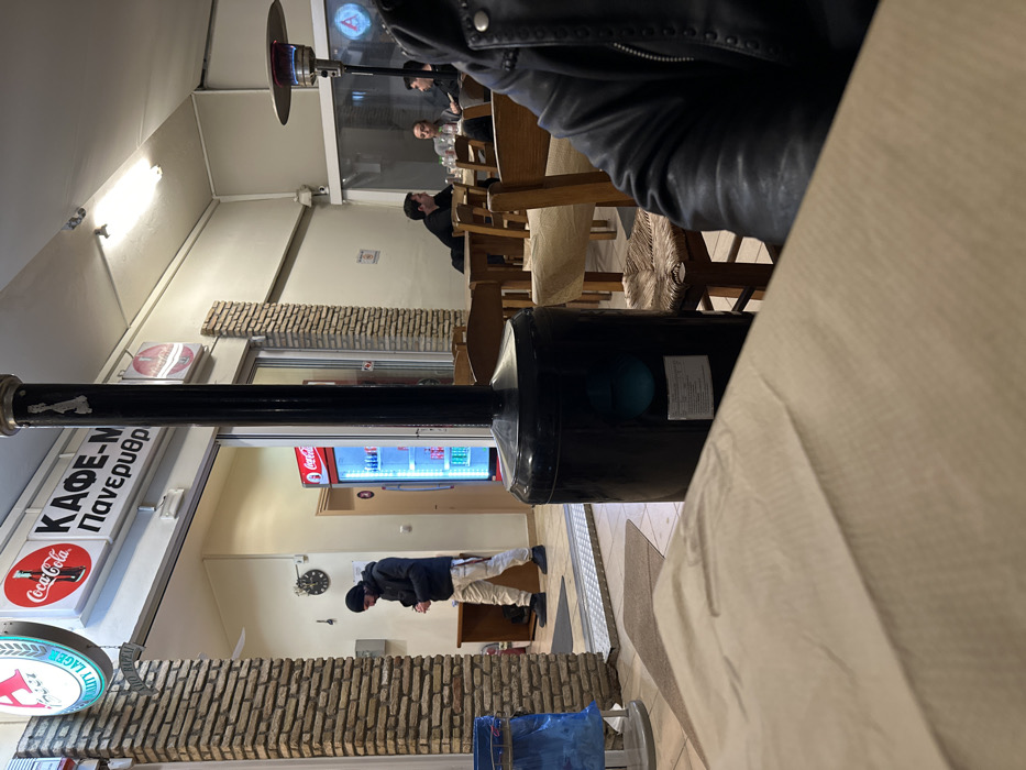 This image captures the interior of what appears to be a casual café or restaurant, likely in Nea Erithrea, Greece, during the evening or night, indicated by the sole reliance on artificial lighting. The shot is taken from a low angle, possibly from a seated position at a table.
In the foreground, the base of a large, black, cylindrical object, potentially an industrial heater or light fixture, partially obscures a beige tabletop. Another tall, silver and black patio heater is visible further back on the left side of the room.
The space is furnished with numerous wooden tables and chairs, some featuring light-colored woven or fabric seat coverings. The walls incorporate decorative brick-patterned sections.
Several individuals are present. One man, dressed in a dark jacket and light trousers with a dark beanie, stands near a glass-fronted Coca-Cola refrigerator filled with drinks. He appears to be looking down at something on a counter or table. Further back, through a large window or opening, two other patrons are seated at a table. Another individual in a dark jacket is seated closer to the foreground, with their back partially turned.
Visible text on signs includes "ΚΑΦΕ-Μ" and "Πανερυθρ" (likely part of "Πανερυθραϊκός," a common local reference in Greece, connecting to the Nea Erithrea context). Below this sign, a circular red Coca-Cola logo is displayed. A partially visible blue and white round sign also reads "QUALITY LAGER." A wall-mounted clock is visible above where the standing man is positioned. The overall atmosphere suggests a relaxed, social setting.