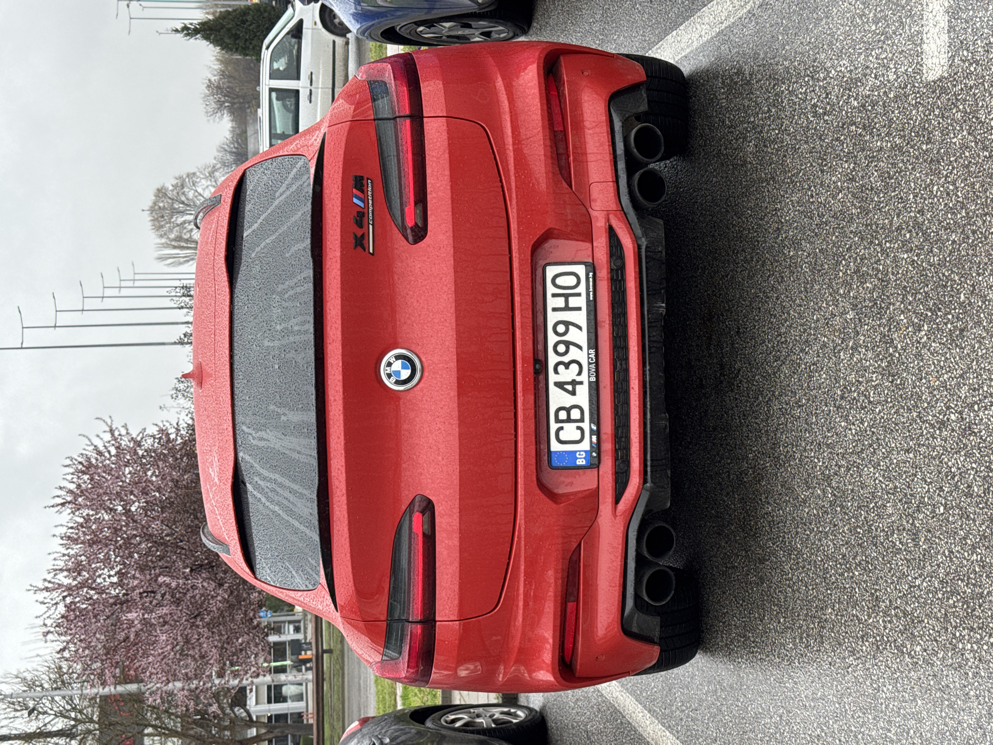The image shows the rear of a red BMW X4 M Competition parked on wet asphalt. The Bulgarian license plate reads "CB 4399 HO". Visible on the car are the BMW logo and the "X4 M Competition" badge. The car has four exhaust pipes, two on each side. The location is likely in Plovdiv, Bulgaria, based on the license plate. A flowering tree is visible in the background, along with other parked cars and street lights. The sky appears overcast.