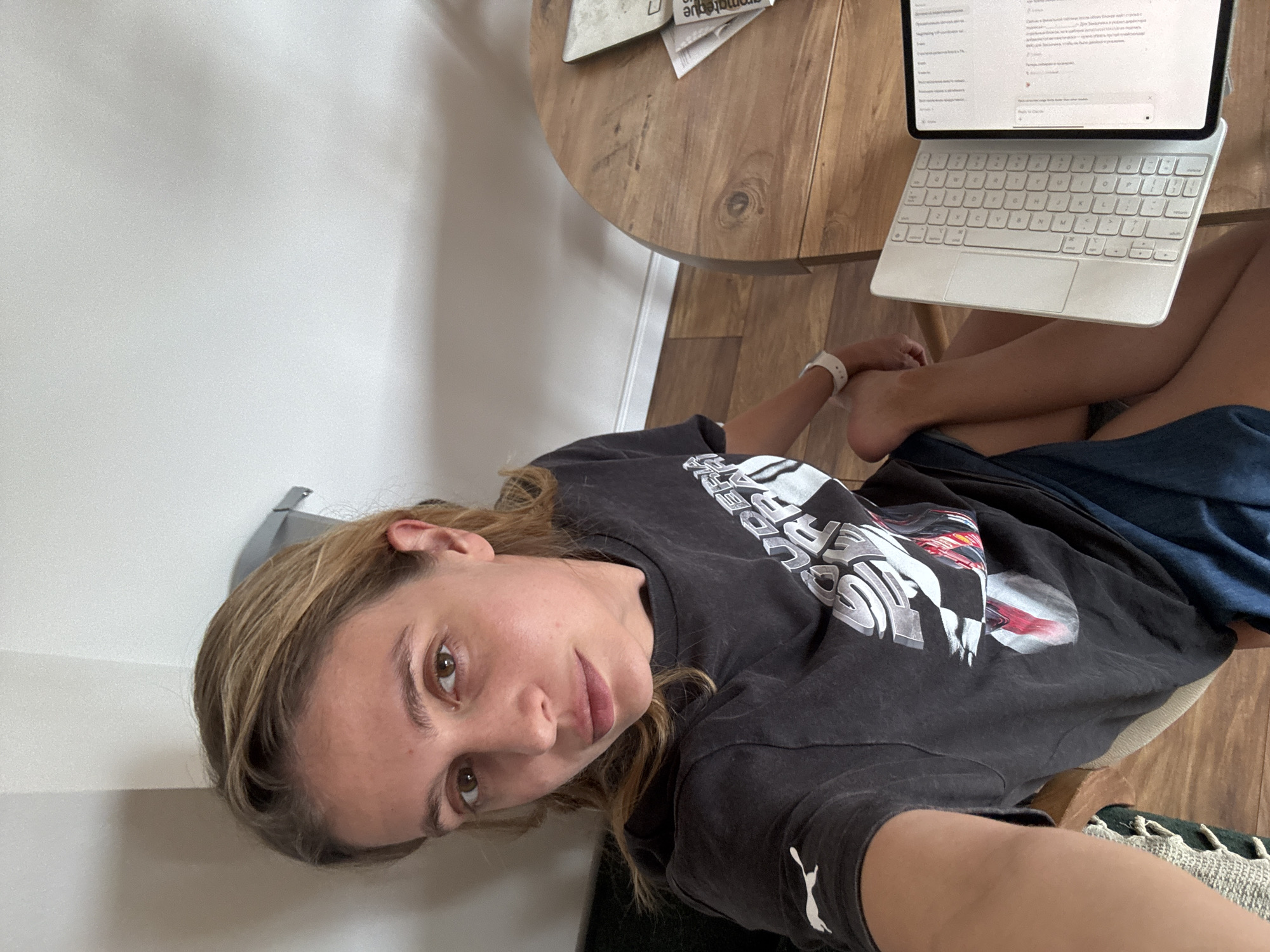 A woman sits at a round wooden table in an indoor environment. A tablet with a keyboard attachment sits on the table, displaying text on its screen. The woman wears a dark Puma T-shirt and blue shorts. She is looking directly at the camera. The background includes a white wall and wooden flooring. The location is Unknown, Unknown.