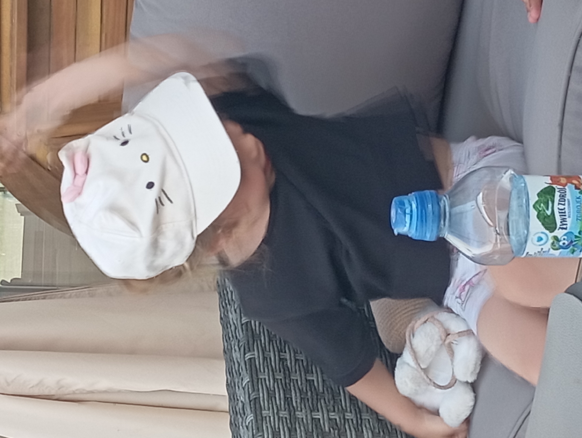 A young child wearing a white cap with a pink bow and Hello Kitty design, and a black t-shirt, is sitting on a grey outdoor sofa. The child is holding a white stuffed animal and has a bottle of water next to them. The background shows a wooden structure and a tan curtain. The scene appears to be in a backyard or patio area.