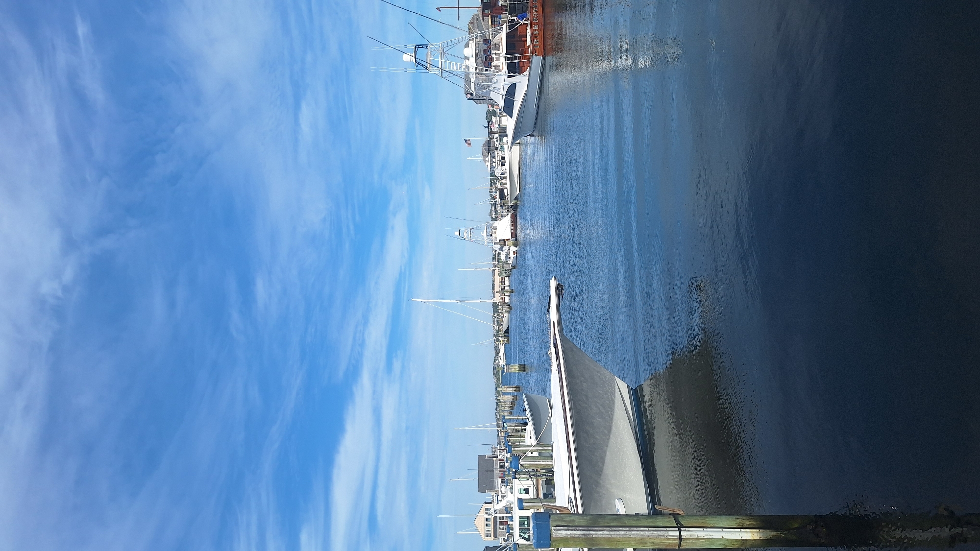 A wide shot shows a harbor in Nantucket, Massachusetts. The water is a deep blue, reflecting the sky. A variety of boats are docked, ranging from small sailboats to larger fishing vessels. The boats are white and gray, with some featuring colorful accents. The sky is a bright blue with wispy white clouds. In the background, a few buildings are visible, suggesting the presence of a town or village. The image is taken from the perspective of someone standing on a dock, with a wooden piling in the foreground. The overall scene is one of a peaceful harbor on a clear day.